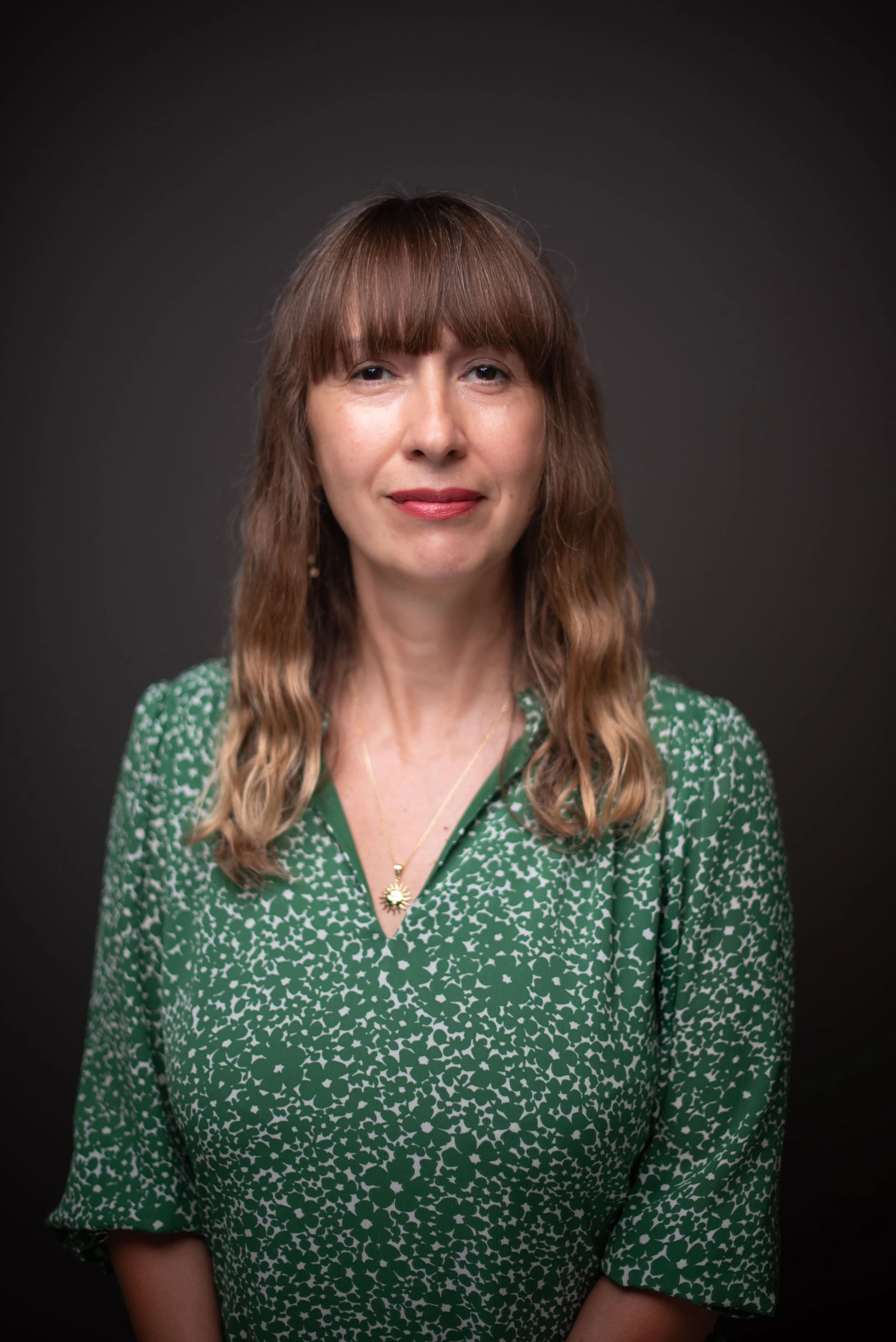 A woman with shoulder-length light brown wavy hair and bangs, wearing a green patterned blouse and gold jewelry, smiling slightly, set against a dark gray background.