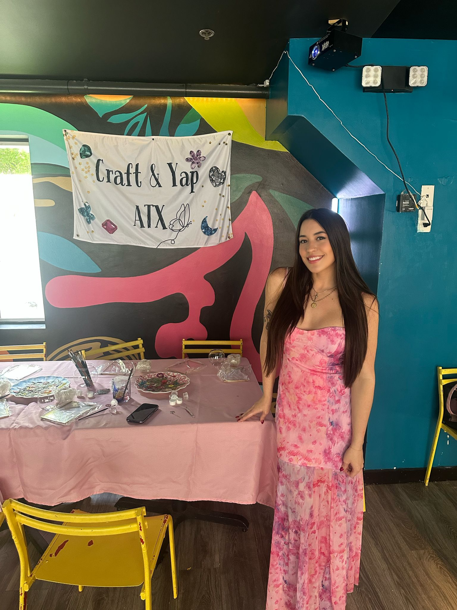 A woman in a pink floral dress standing next to a decorated table with pink tablecloth, plates, and crafting supplies, at a craft and yap event in Austin, Texas, with a colorful mural in the background and a sign that reads 'Craft & Yap ATX'.