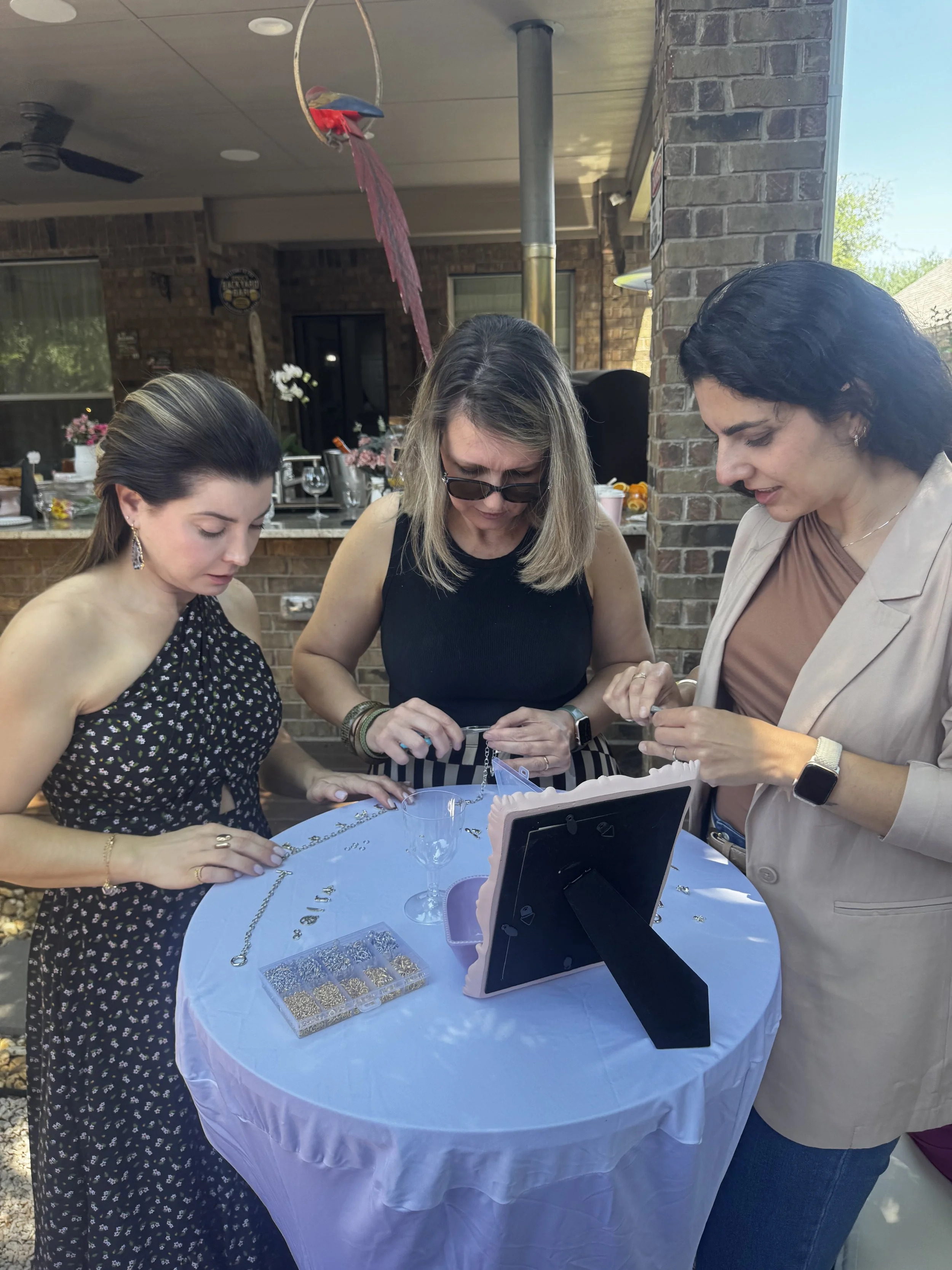 Three women examining jewelry on a round table at an outdoor gathering.