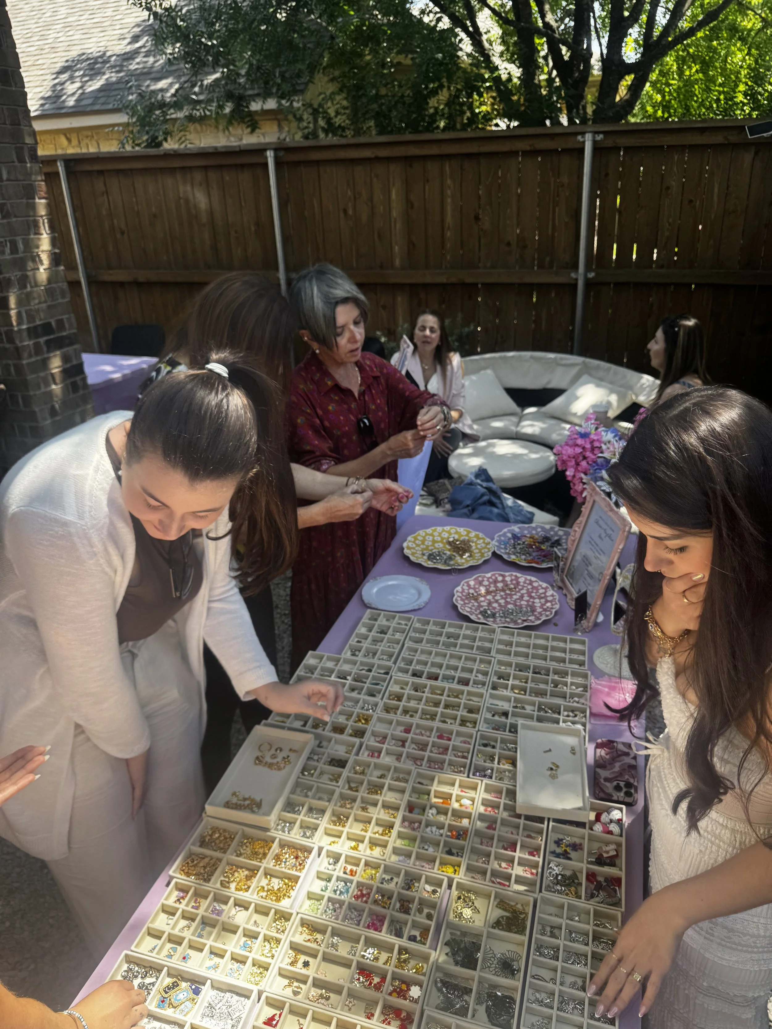 People browsing jewelry and accessories at an outdoor craft fair or market, with a table displaying trays filled with various earrings, brooches, and other jewelry.