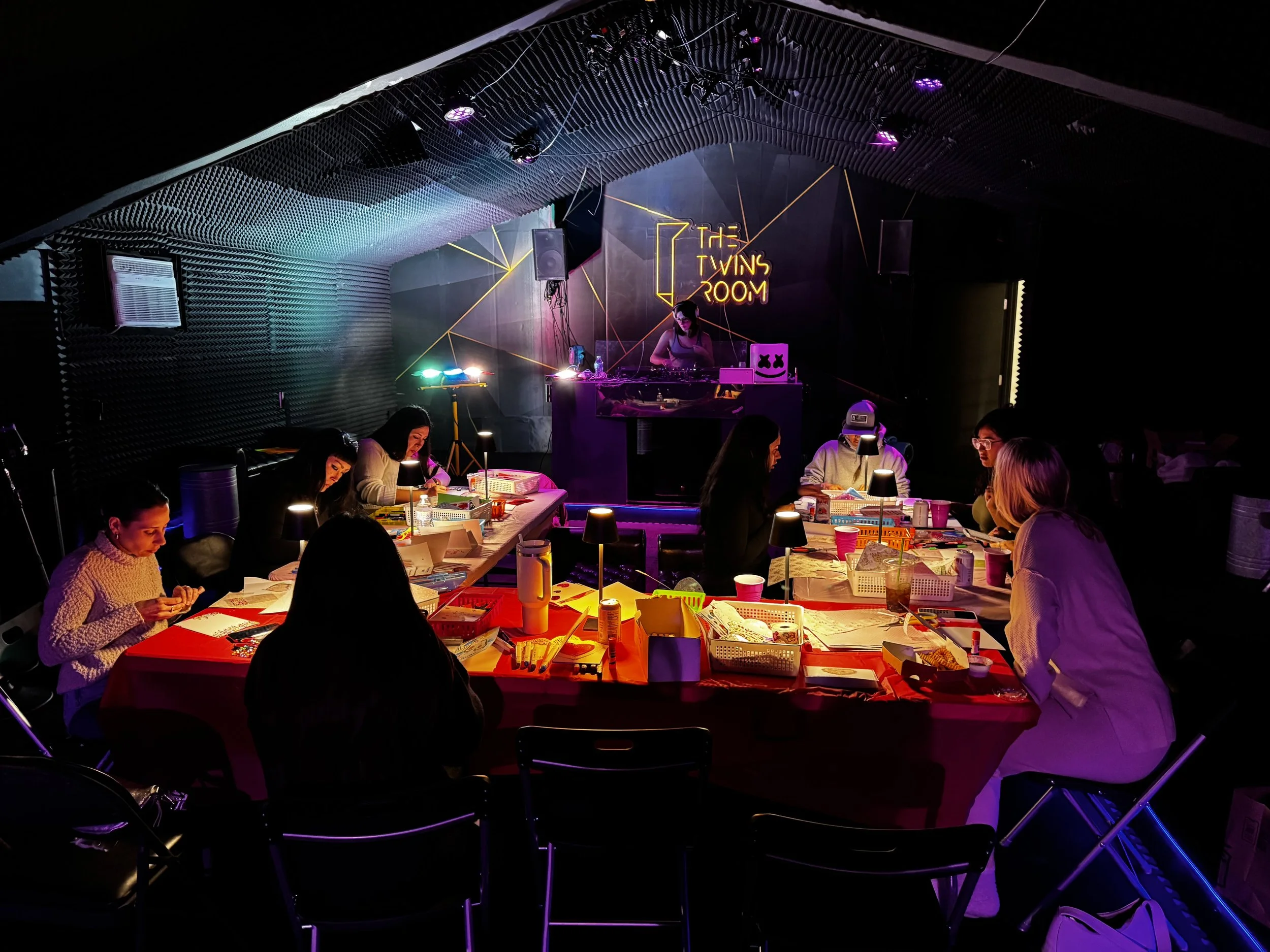 People sitting at tables in a dimly lit room with a DJ on stage, neon sign reading 'The Twins Room', and colorful lights, engaging in arts and crafts.