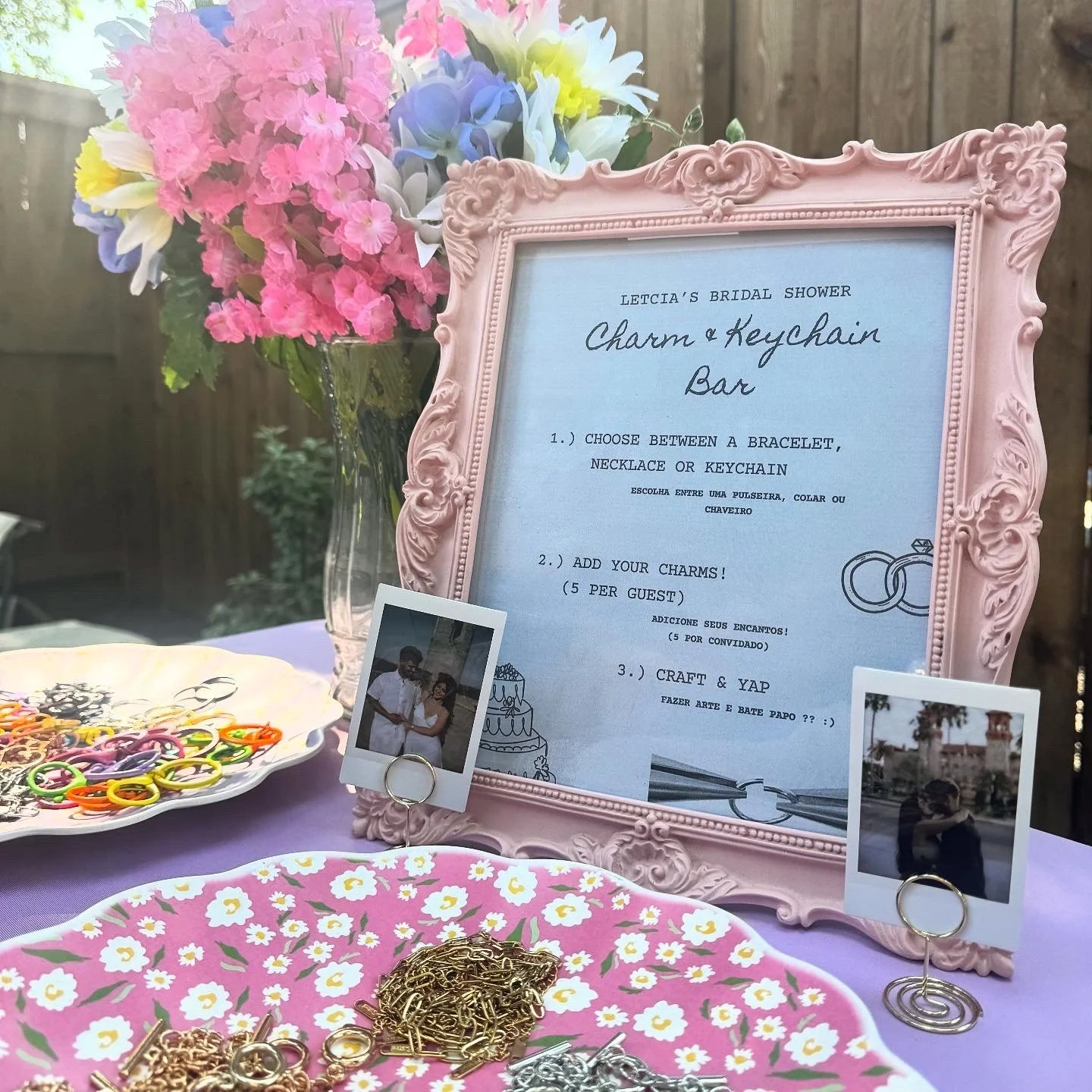 A bridal shower table setup with a pink floral bouquet, a pink ornate picture frame with a schedule for a charm and keychain bar, and two small photograph clips with images of people. There are colorful rubber bands and gold jewelry on a pink floral tray.