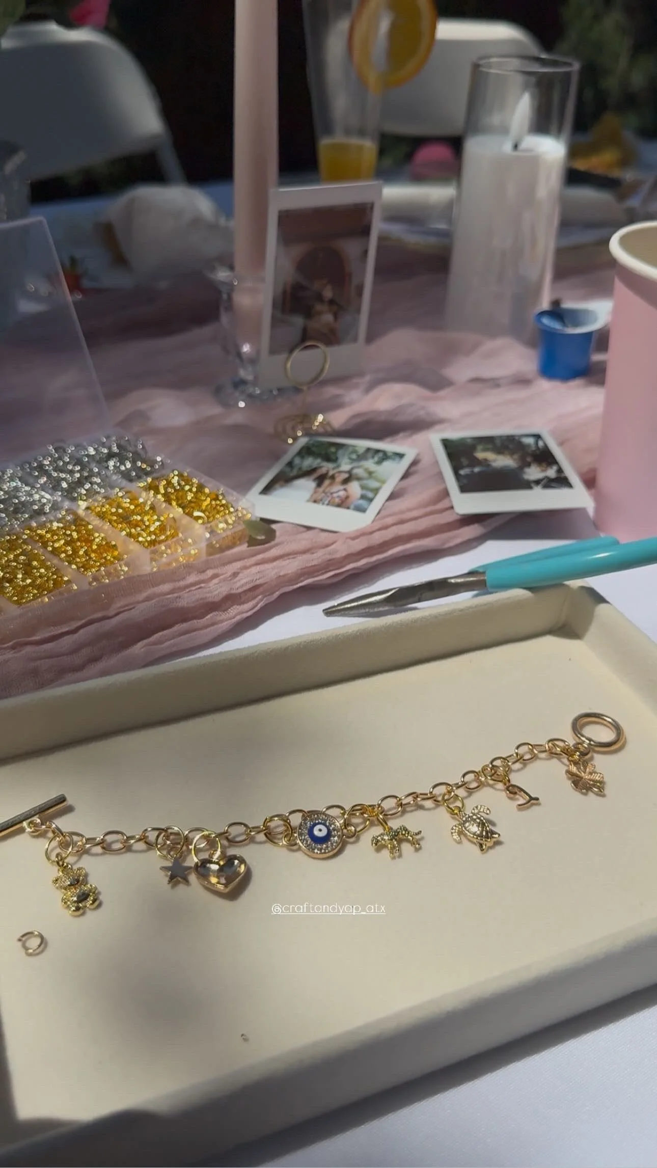 A gold charm bracelet with various charms, including a bee, star, heart, evil eye, camel, and horse, displayed in a jewelry box on a table. The table has a pink cloth with photo prints, a blue pen, and beads. In the background, there are glasses, a candle, and a lemon slice.