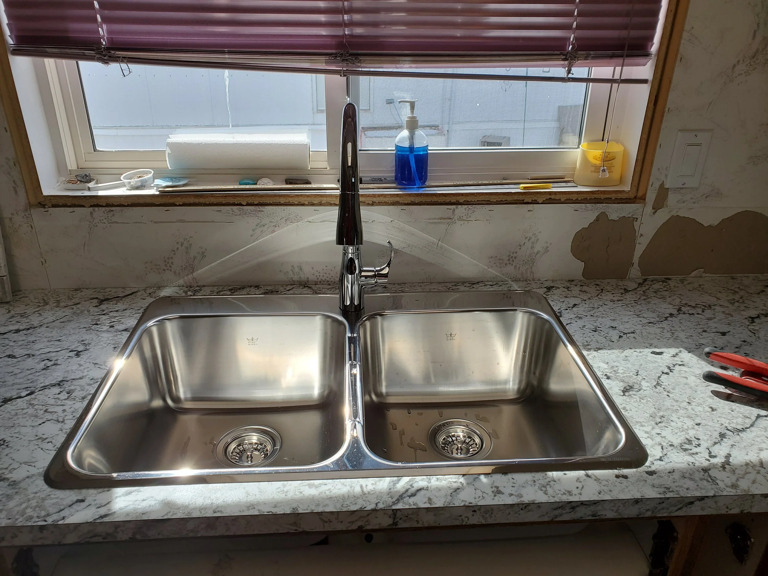 Kitchen sink with a multiple faucet on a marble countertop, window with blinds, soap dispenser, and some small items on the window sill.
