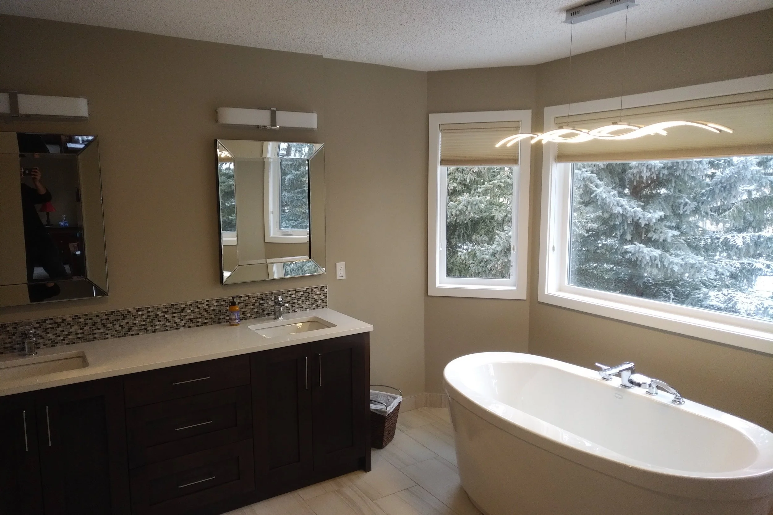 Modern bathroom with a freestanding bathtub, two large windows showing snow-covered trees, a dark wood vanity with dual sinks and a mosaic tile backsplash, mirror cabinets, and a sleek ceiling light fixture.