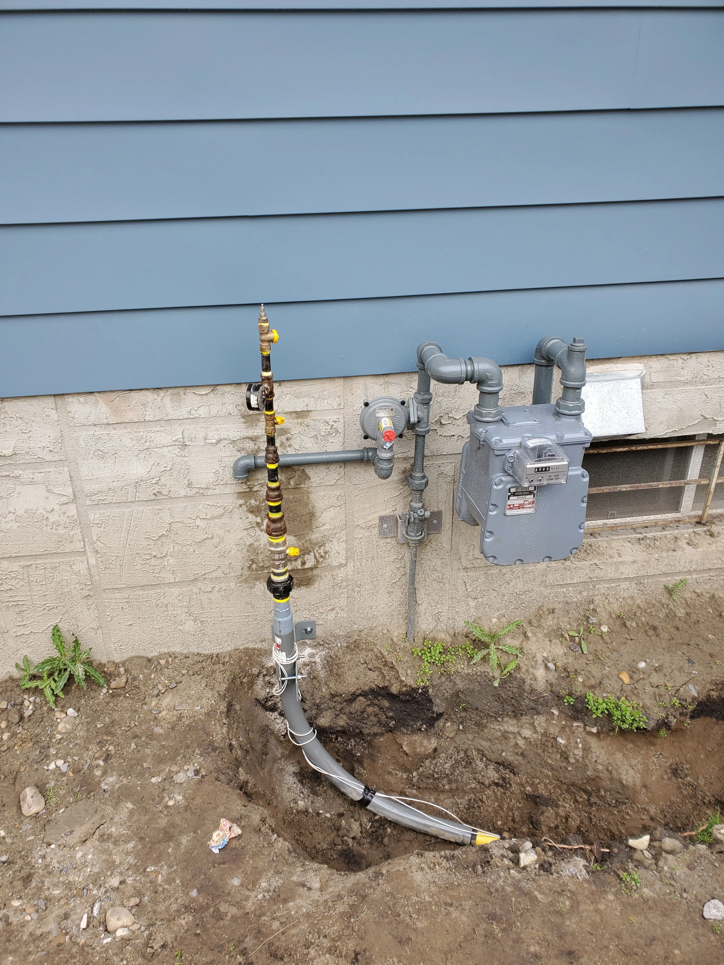 Underground utility meter and piping installed outside a house, with a trench dug for the pipe.