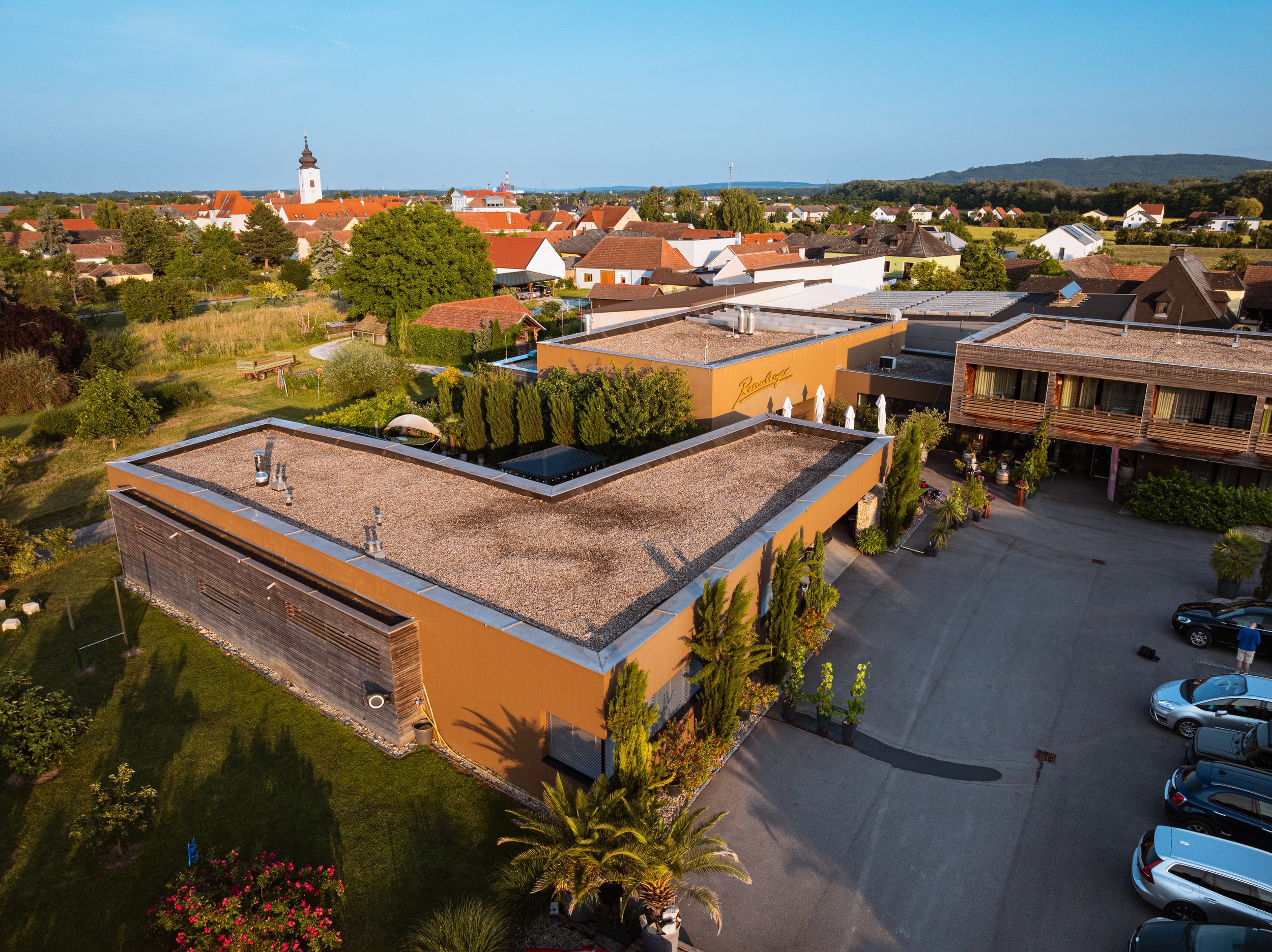 Vogelperspektive auf ein modernes Hotel mit Parking und Garten, umgeben von einem Dorf mit Kirchturm und Hügel im Hintergrund.