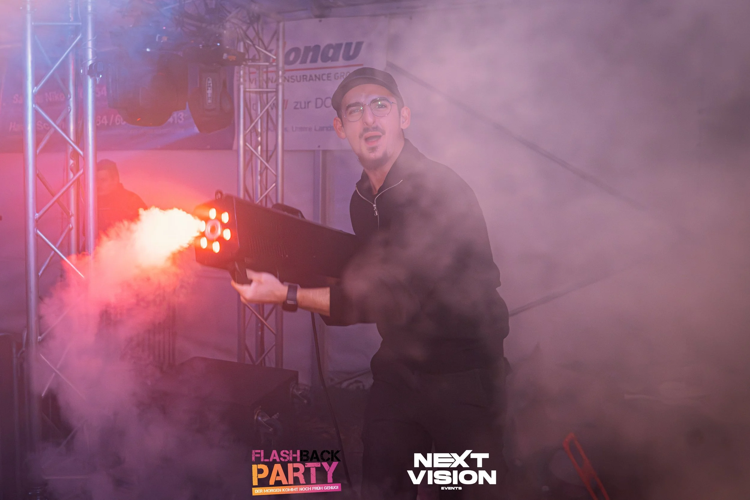Ein junger Mann trägt eine schwarze Jacke, eine schwarze Mütze und runde Brillen. Er hält eine Feuerwaffe, aus der Feuer und Rauch kommen, in einer Nebel- oder Rauchumgebung bei einer Veranstaltung, mit technischen Bühnenstrukturen im Hintergrund.