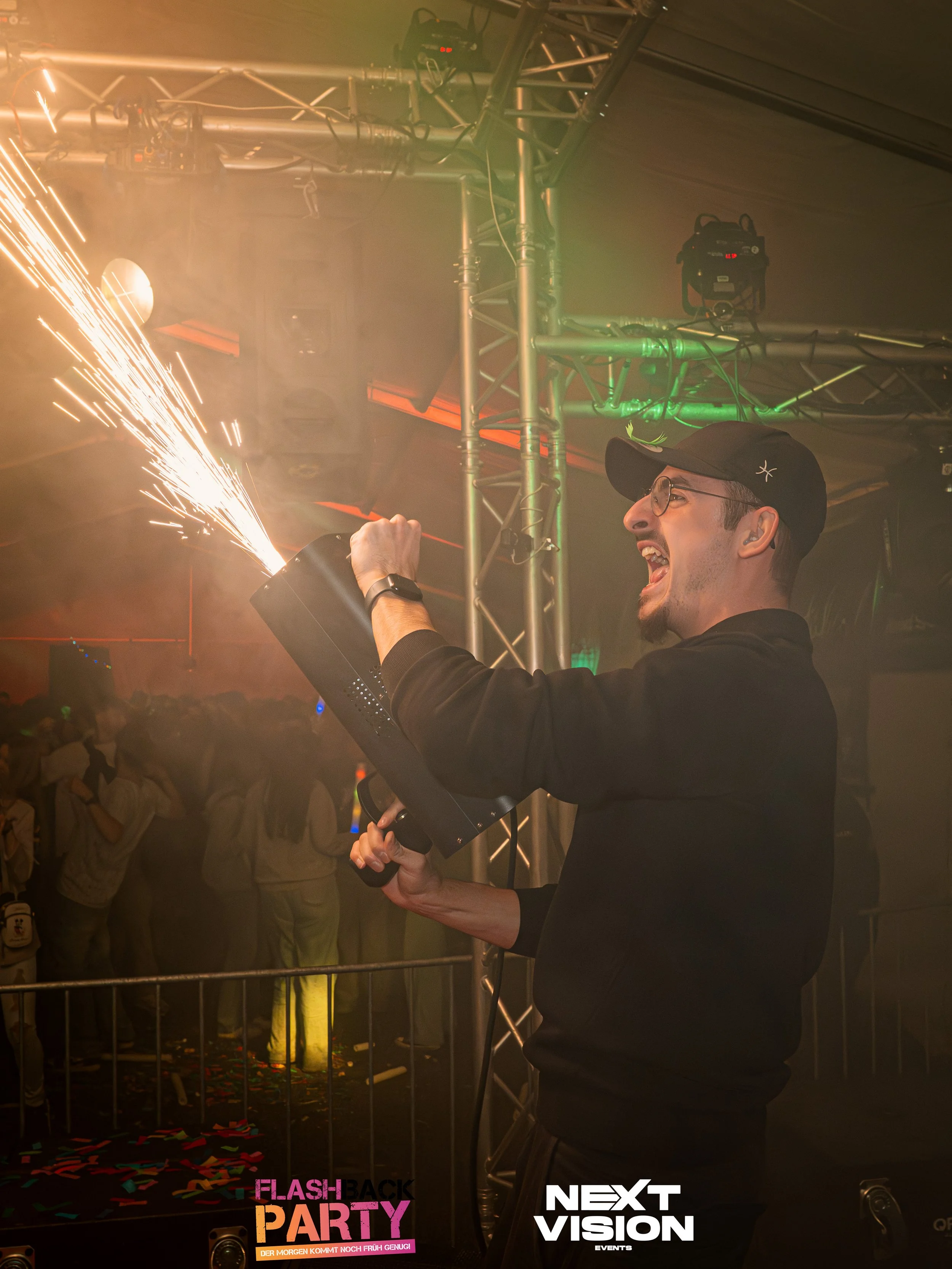 Ein junger Mann mit Brille und schwarzem Hut hält eine mit Funken schießernde Maschine auf einer Party, im Hintergrund tanzen Menschen.