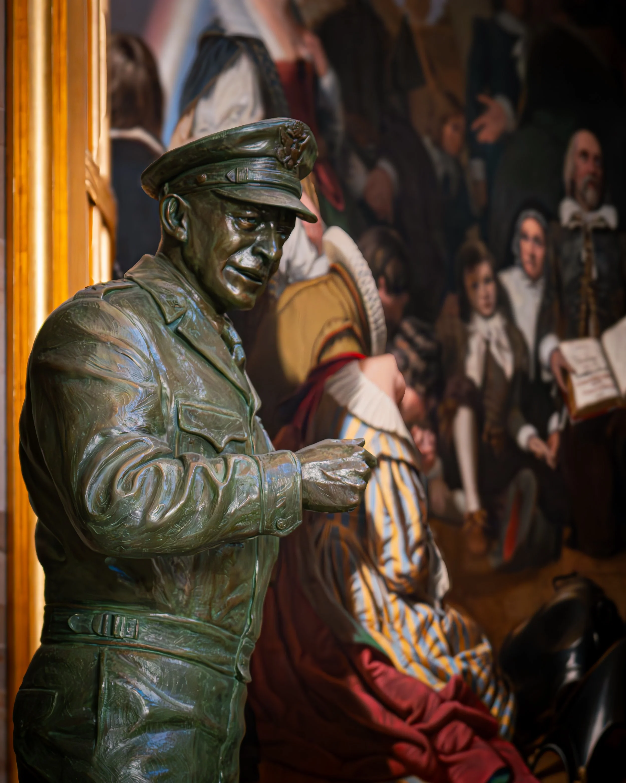 Bronzefigur eines Mannes in Uniform, umgeben von einer Wand mit Gemälden, die Menschen in historischem Gewand zeigen.