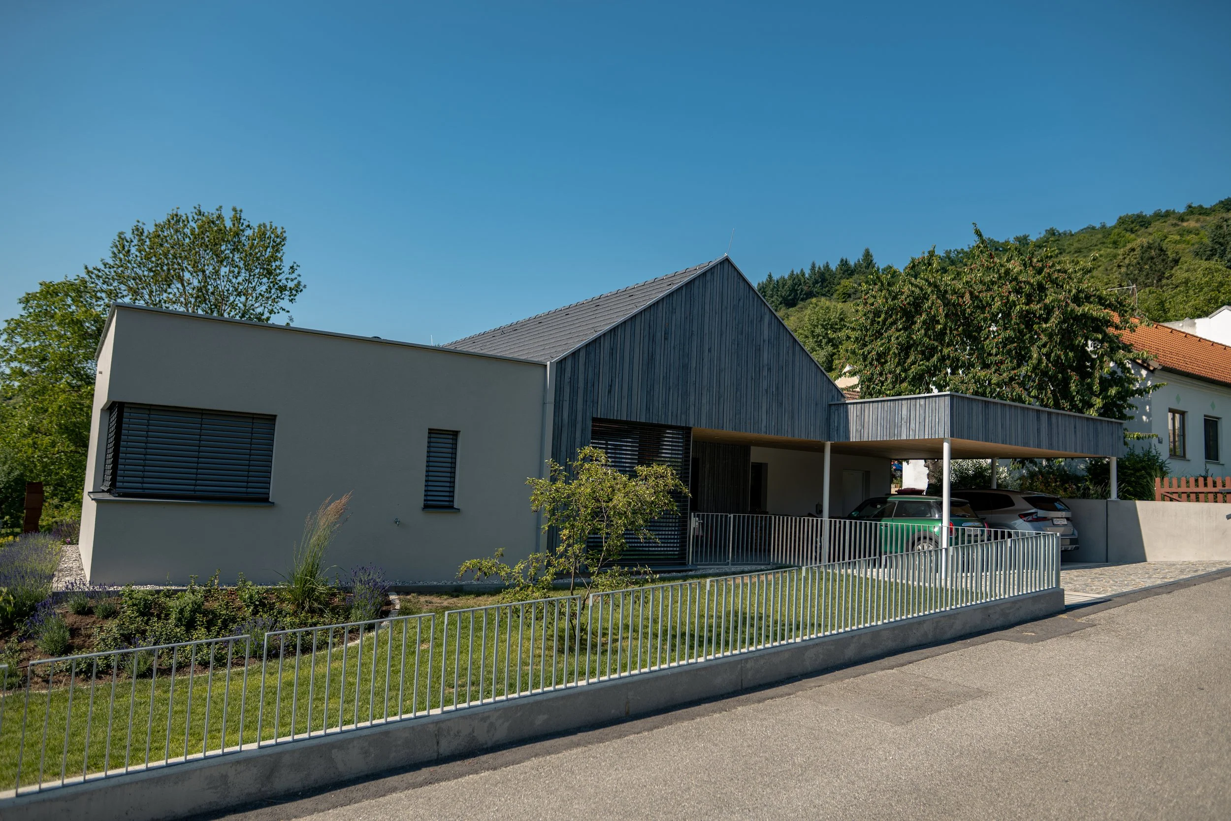 Modernes, zweistöckiges Haus mit weißer und grauer Fassade, Carport mit drei Autos, umgeben von einem gepflegten Garten und grünen Bäumen, im Hintergrund ein grüner Hügel.