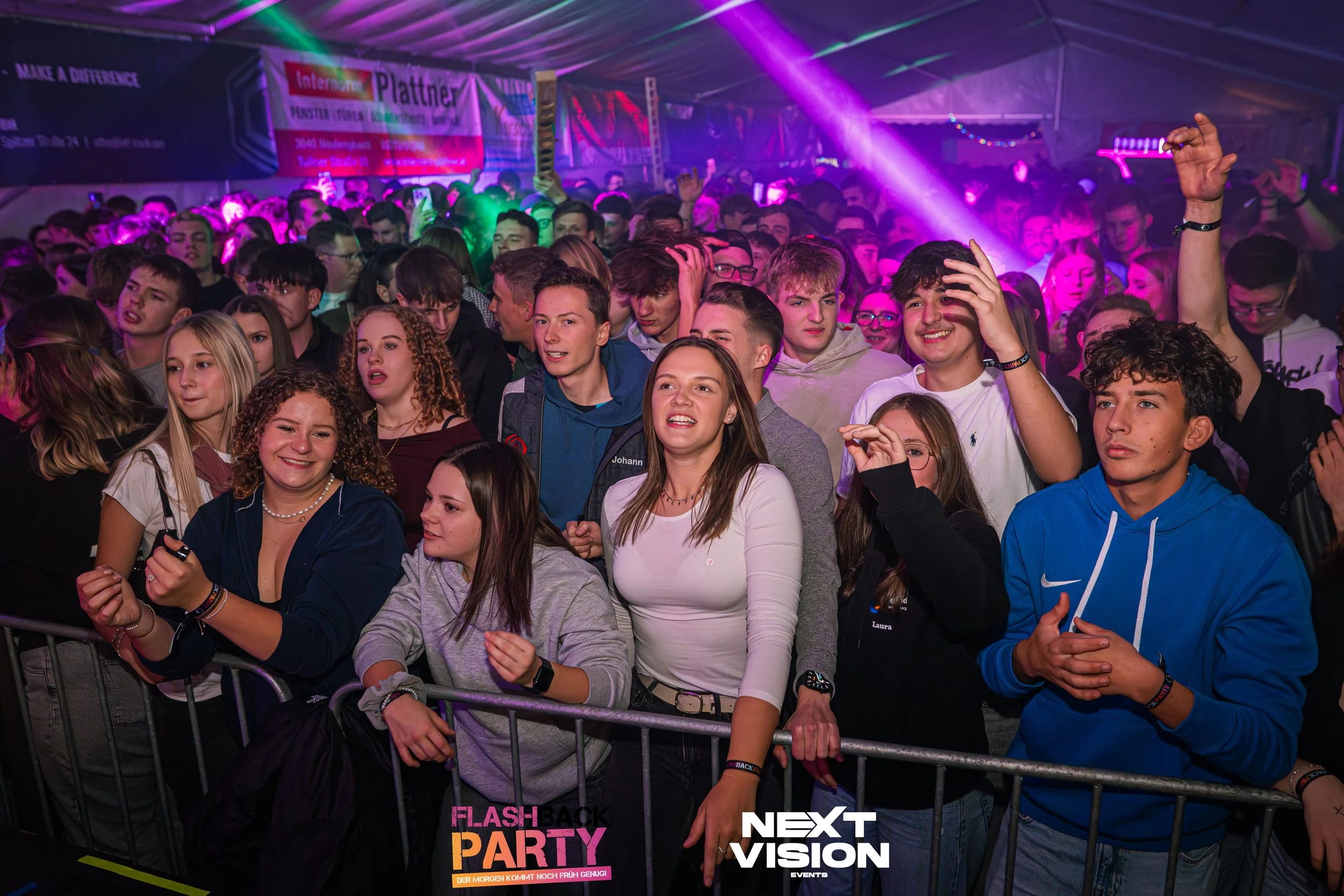Junge Menschen auf einer Partystimmung im Inneren eines Zeltes mit bunten Lichtern, bannerartige Dekorationen und einer Bar im Hintergrund, die bei einer Tanzparty teilnehmen.