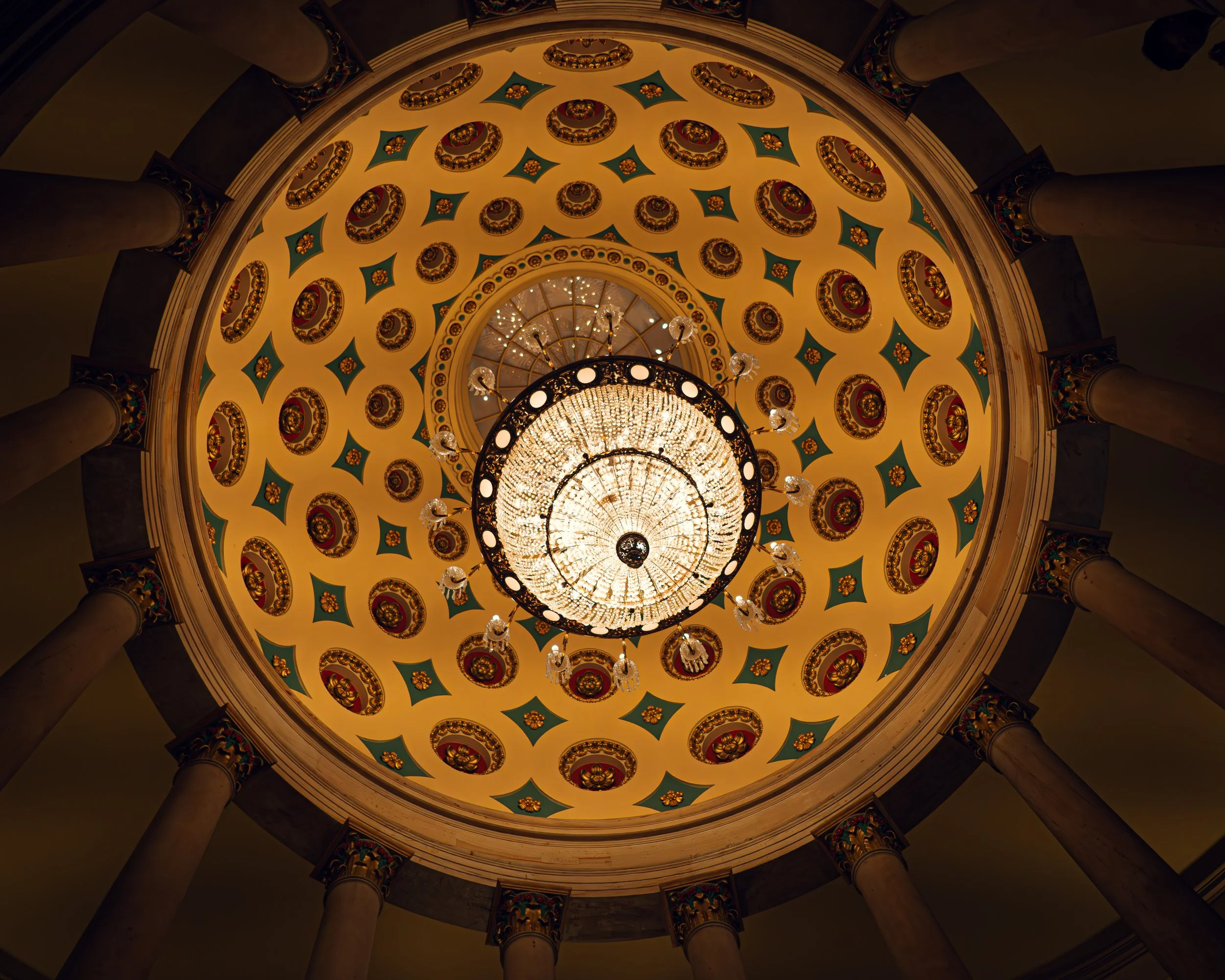 Blick nach oben in eine verzierten Decke mit einer großen, leuchtenden Kronleuchter in einem historischen Gebäude, umgeben von Säulen.