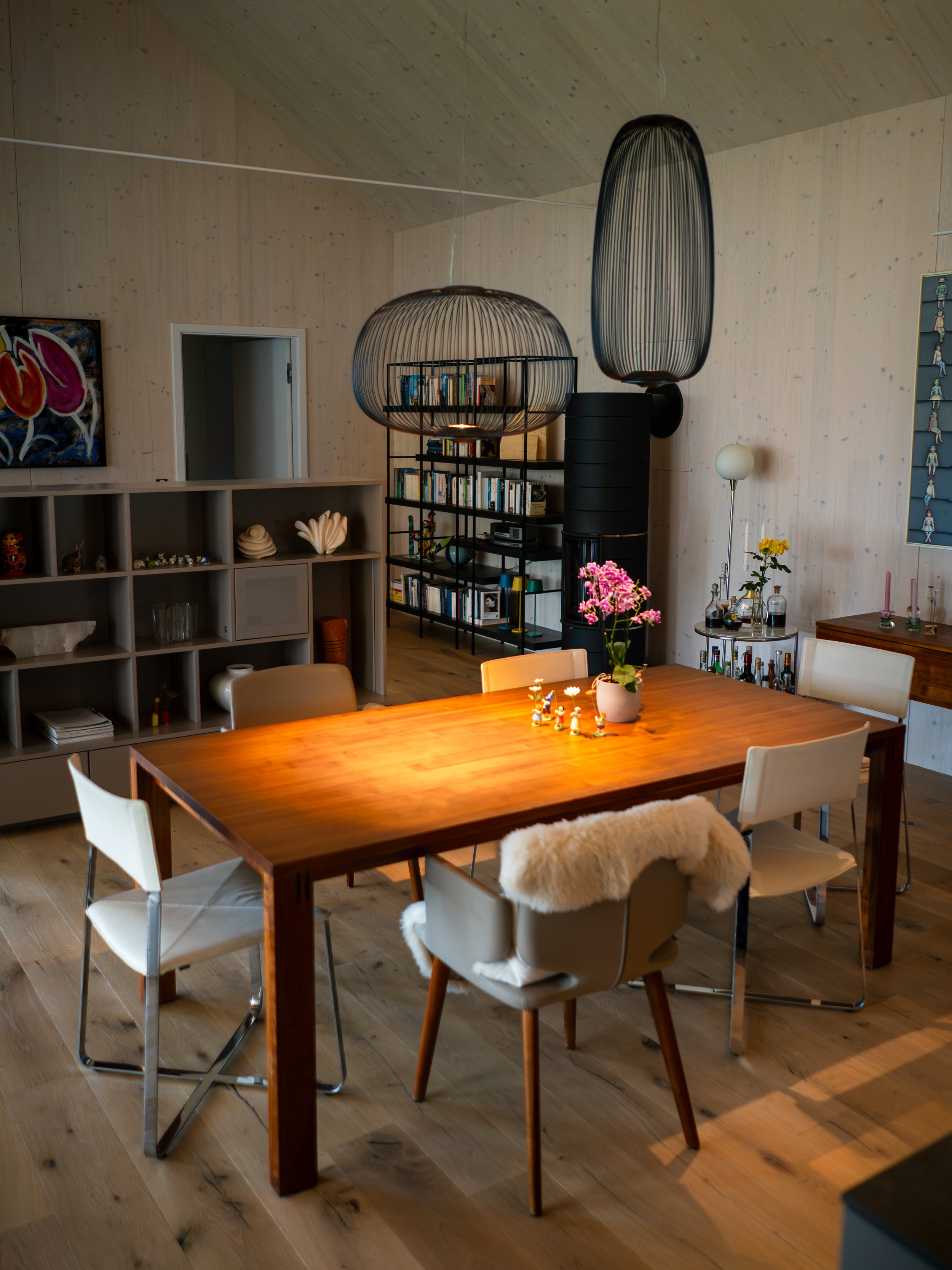 Ein modern eingerichteter Essbereich mit einem Holztisch, sechs Stühlen, einer Topfpflanze im Mittelpunkt. Im Hintergrund sind Bücherregale, dekorative Gegenstände, eine gläserne Lampe und eine schwarze Kaminanlage. Die Wände sind in hellem Holz geha