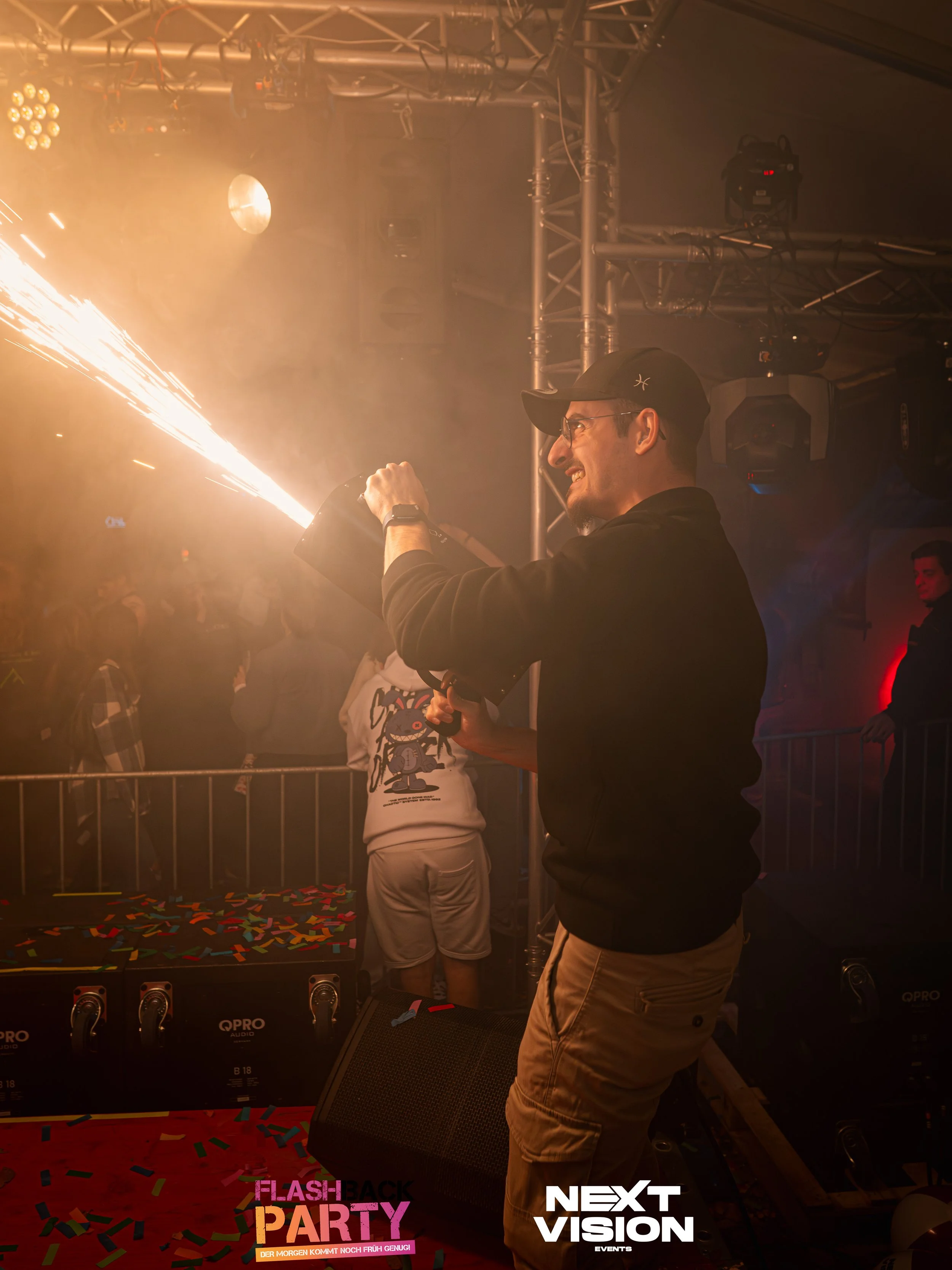 Ein Mann mit Brille und schwarzer Jacke hält eine Feuerwerksrakete in der Hand und lässt sie explodieren, während er auf einer Party ist. Im Hintergrund sind Menschen und Lichteffekte zu sehen, auf dem Boden liegen bunte Konfettistücke.