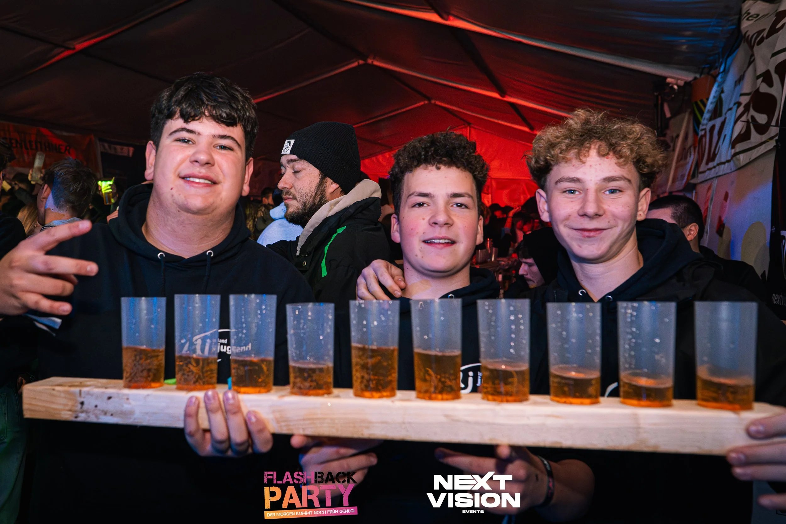 Drei junge Männer auf einer Party, die ein Tablett mit mehreren Biergläsern halten, freundschaftlich lachend, im Hintergrund weitere Partygäste und bunte Beleuchtung.