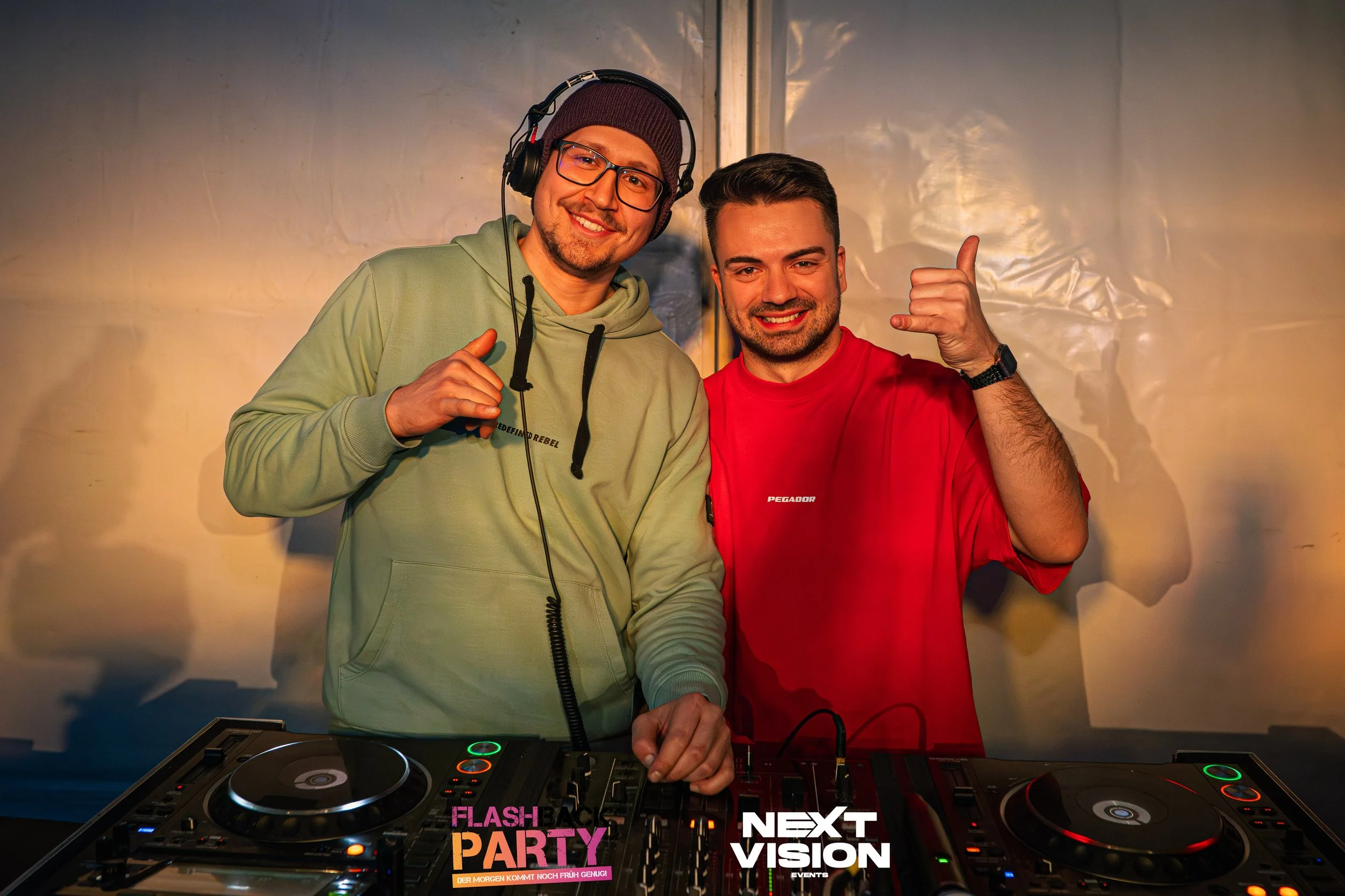 Zwei junge Männer stehen vor DJ-Equipment im Partyraum, lachen und zeigen Daumen hoch. Einer trägt Kopfhörer, beide sind in Freizeitkleidung. Der Hintergrund ist eine graue Wand mit Lichtreflexionen.