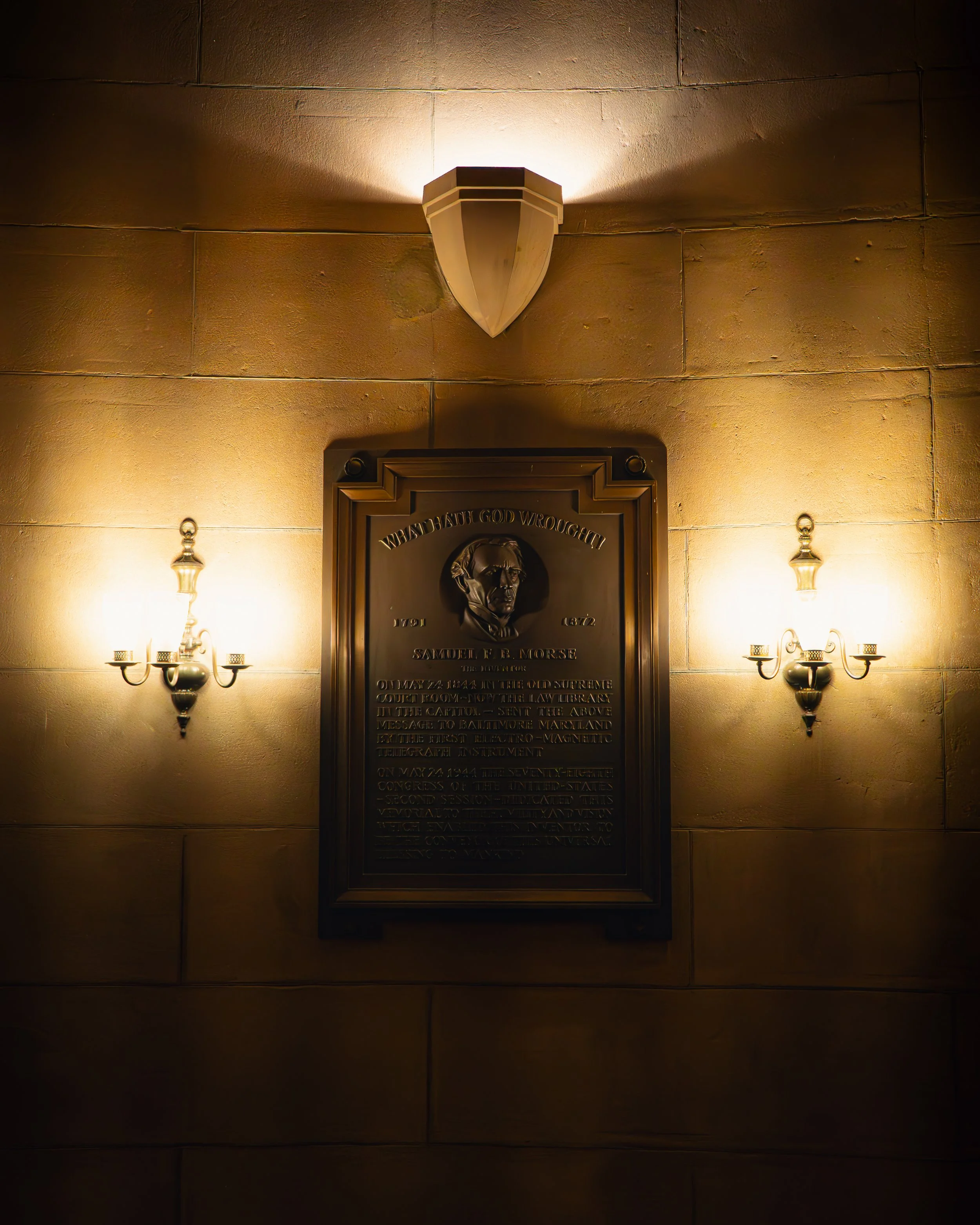 Hinweisschild mit einem Porträt von Samuel F. B. Morse, beleuchtet an einer Wand mit zwei Wandleuchten.