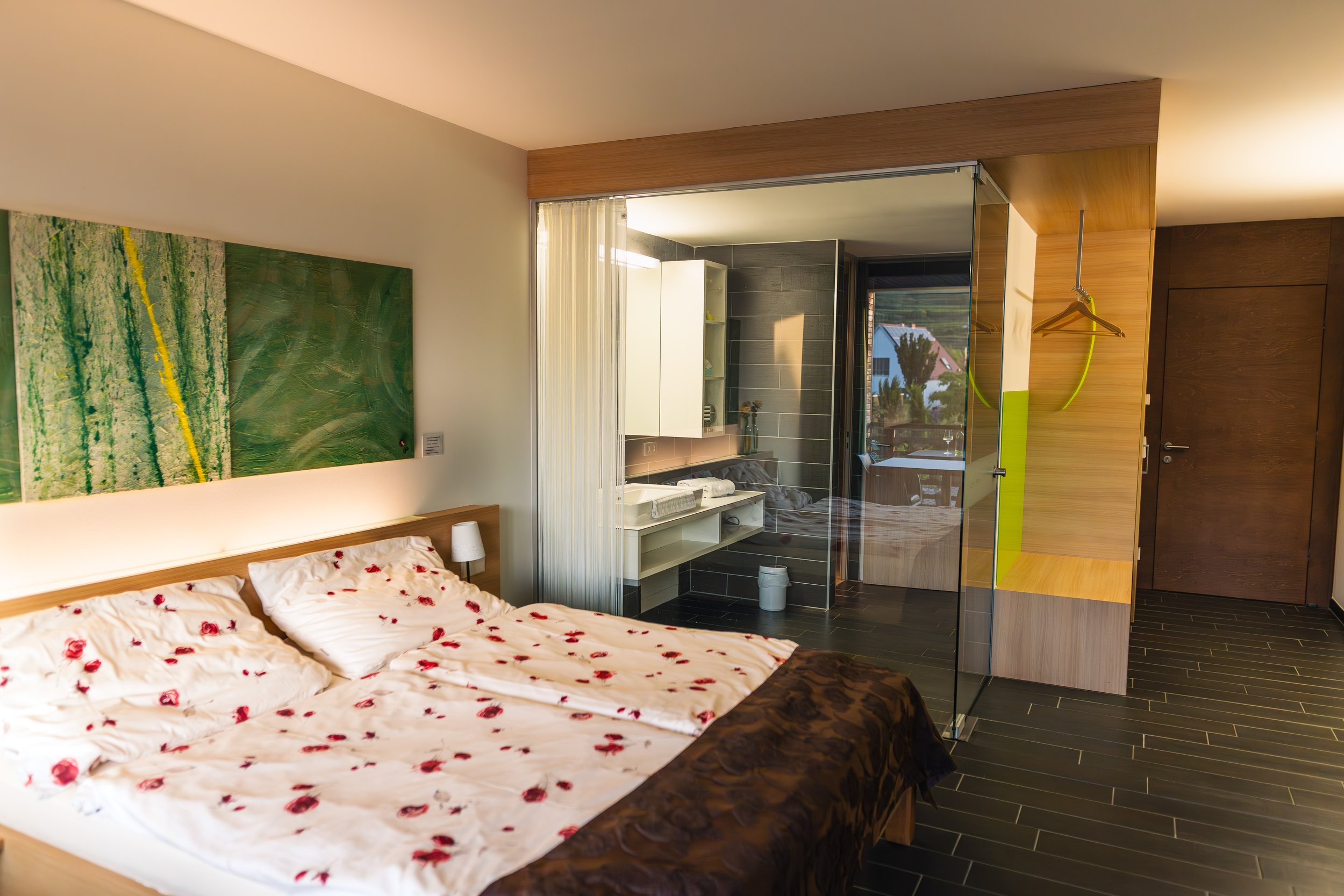 Modernes Hotelzimmer mit Doppelbett, das mit Rosenblättern bedeckt ist, und offenem Badbereich mit Waschbecken, Dusche und Balkon mit Blick ins Freie.