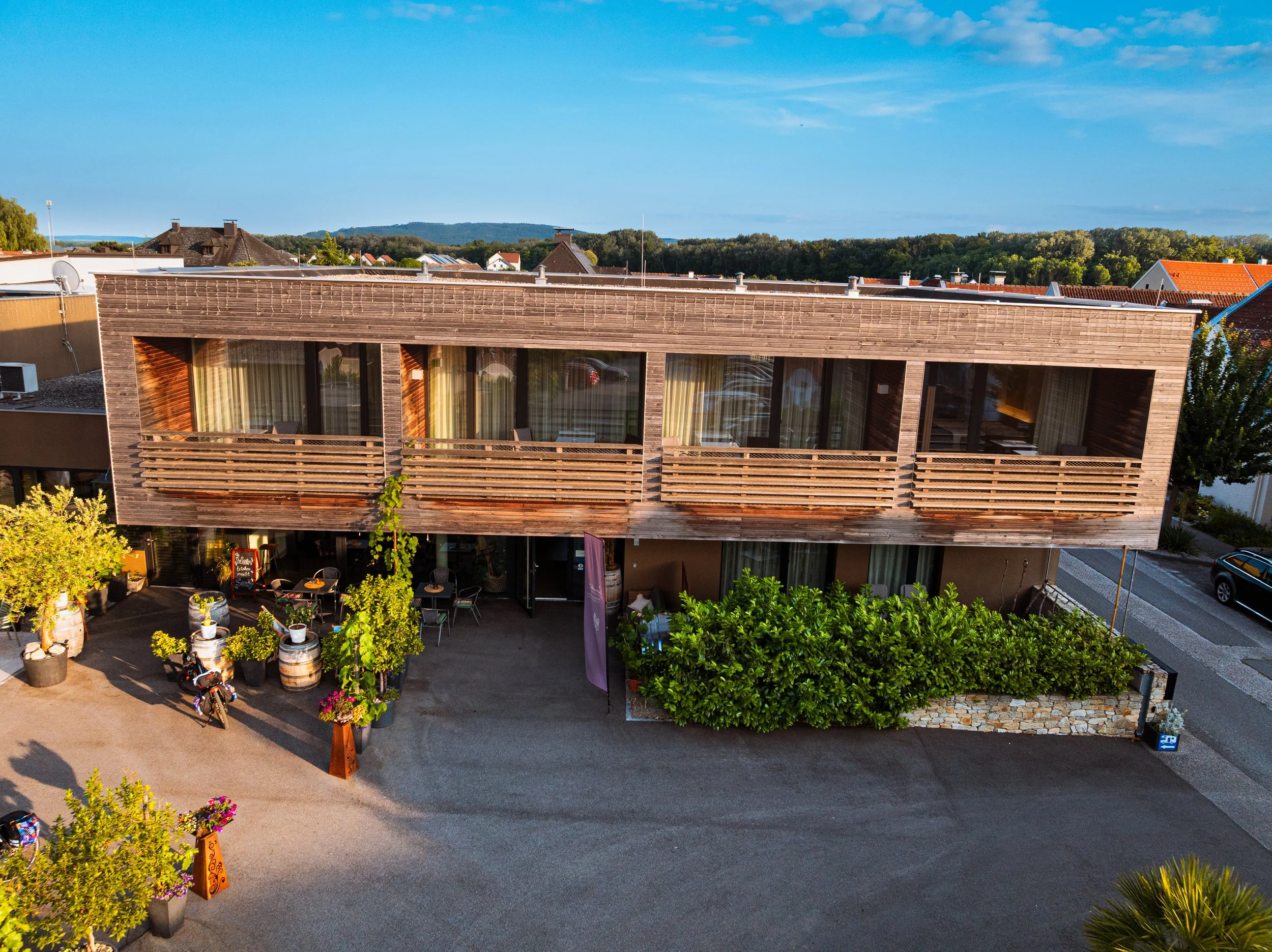 Ein modernes, zweistöckiges Gebäude mit Holzfassade und Balkon, umgeben von Pflanzen, Autos und einem Parkplatz, bei gutem Wetter.