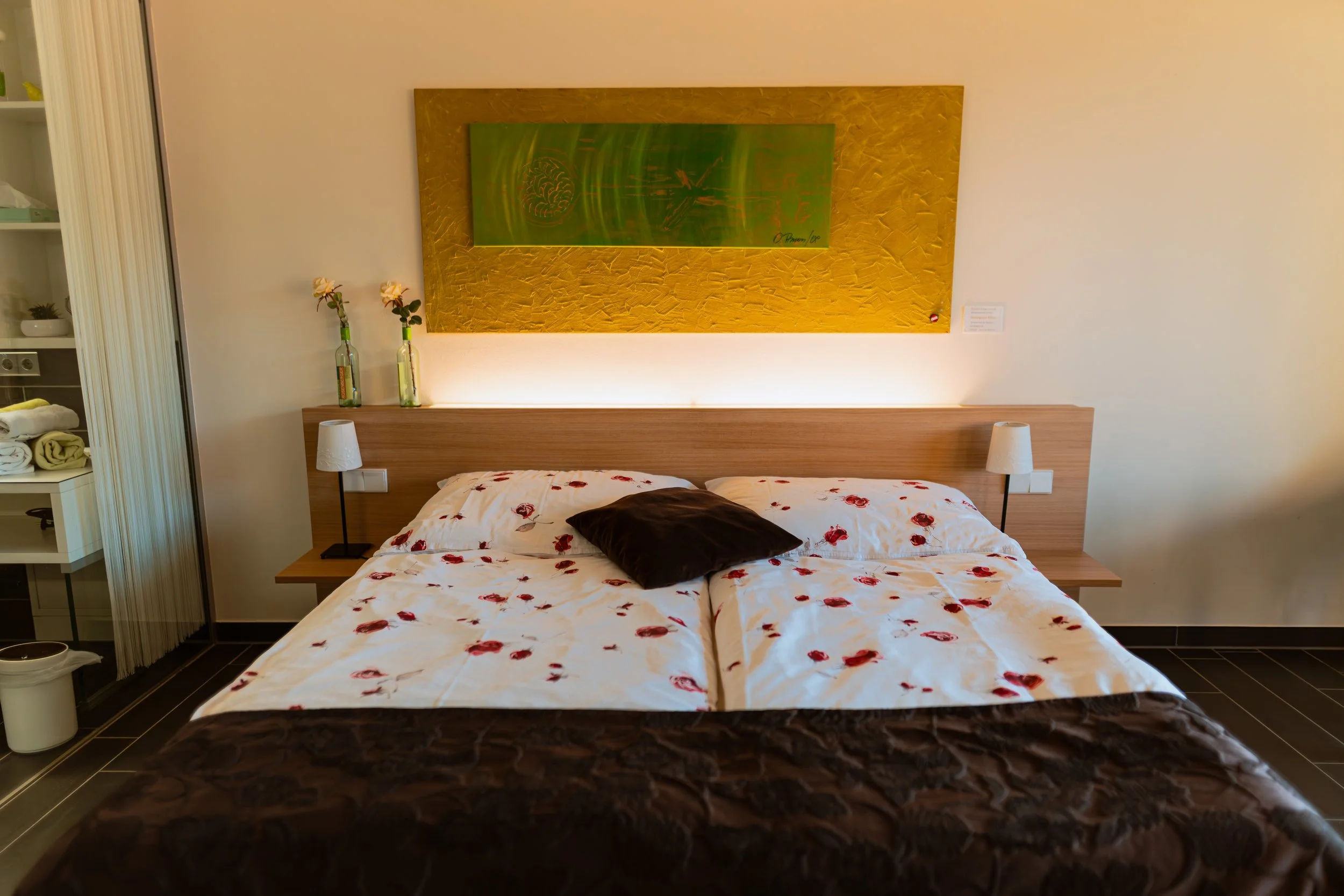 Ein Bett mit weiße Bettwäsche, die mit roten Blüten bedruckt ist, und einem schwarzen Kissen in einem Hotelzimmer. Es gibt zwei Nachttische mit kleinen Lampen, eine Wanddekoration über dem Bett und eine offene Schränkecke auf der linken Seite.