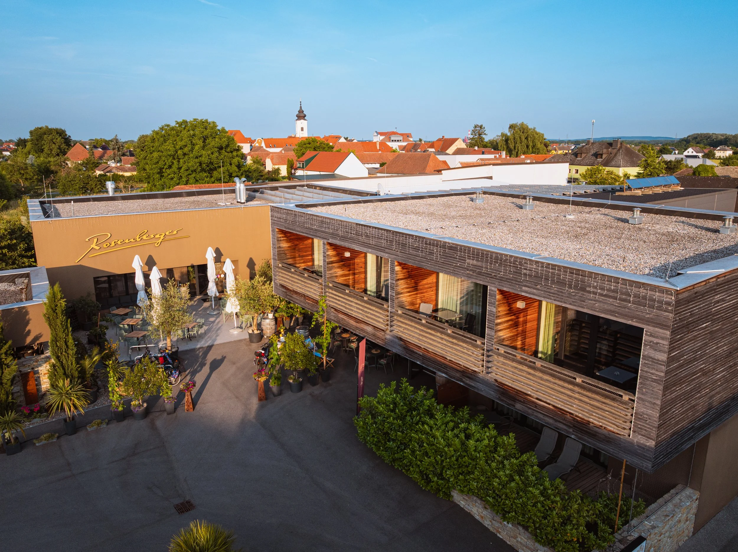 Außenansicht eines modernen Restaurants oder Cafés mit Terrassenbereich, Pflanzen und Tischen im Vordergrund, und einem Wohngebiet mit Kirchturm im Hintergrund.