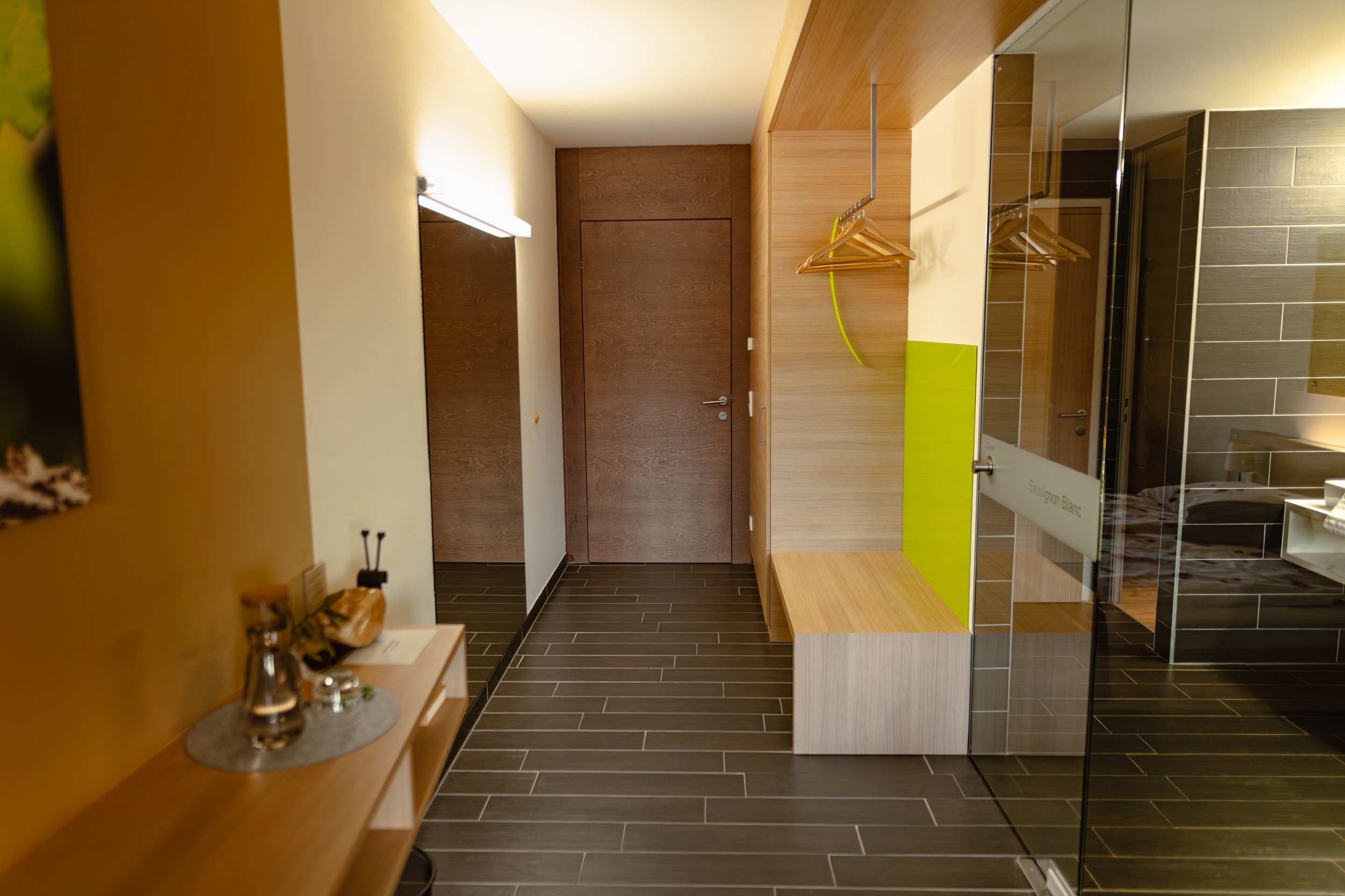 Innenraum eines modern eingerichteten Hotelzimmers mit Flur, Holzlampe, Spiegel, Holzbank, Kleiderhaken, Dusche mit Glastür, grauer Fliesenboden.