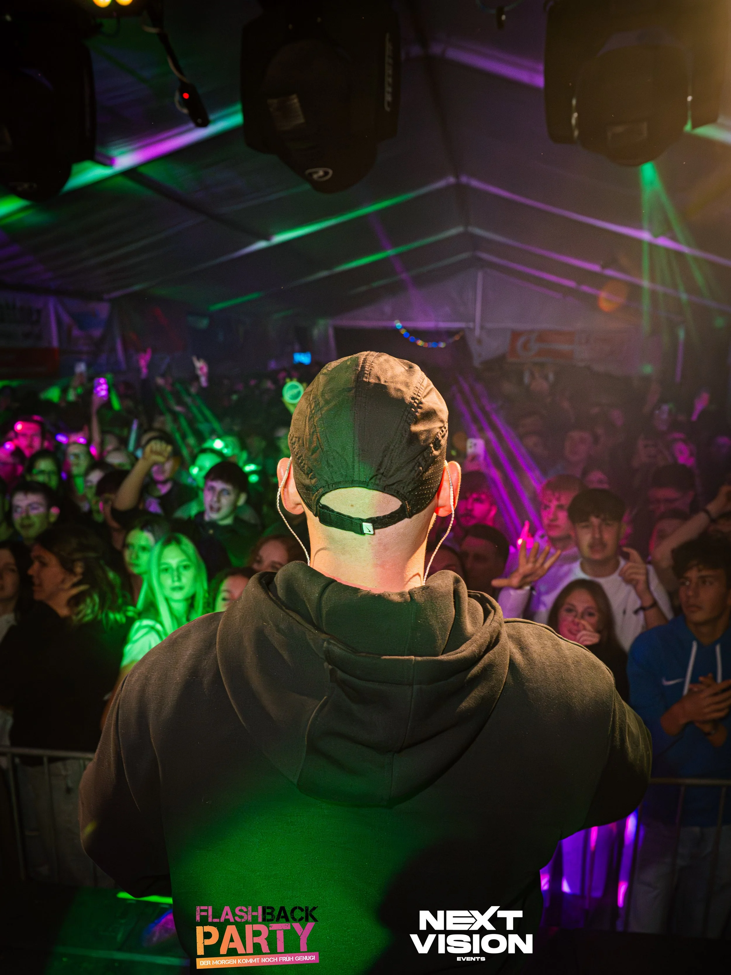 Ein DJ oder Veranstalter blickt auf eine tanzende Menge bei einer Party, farbenfrohe Lichter und Lasereffekte im Hintergrund.