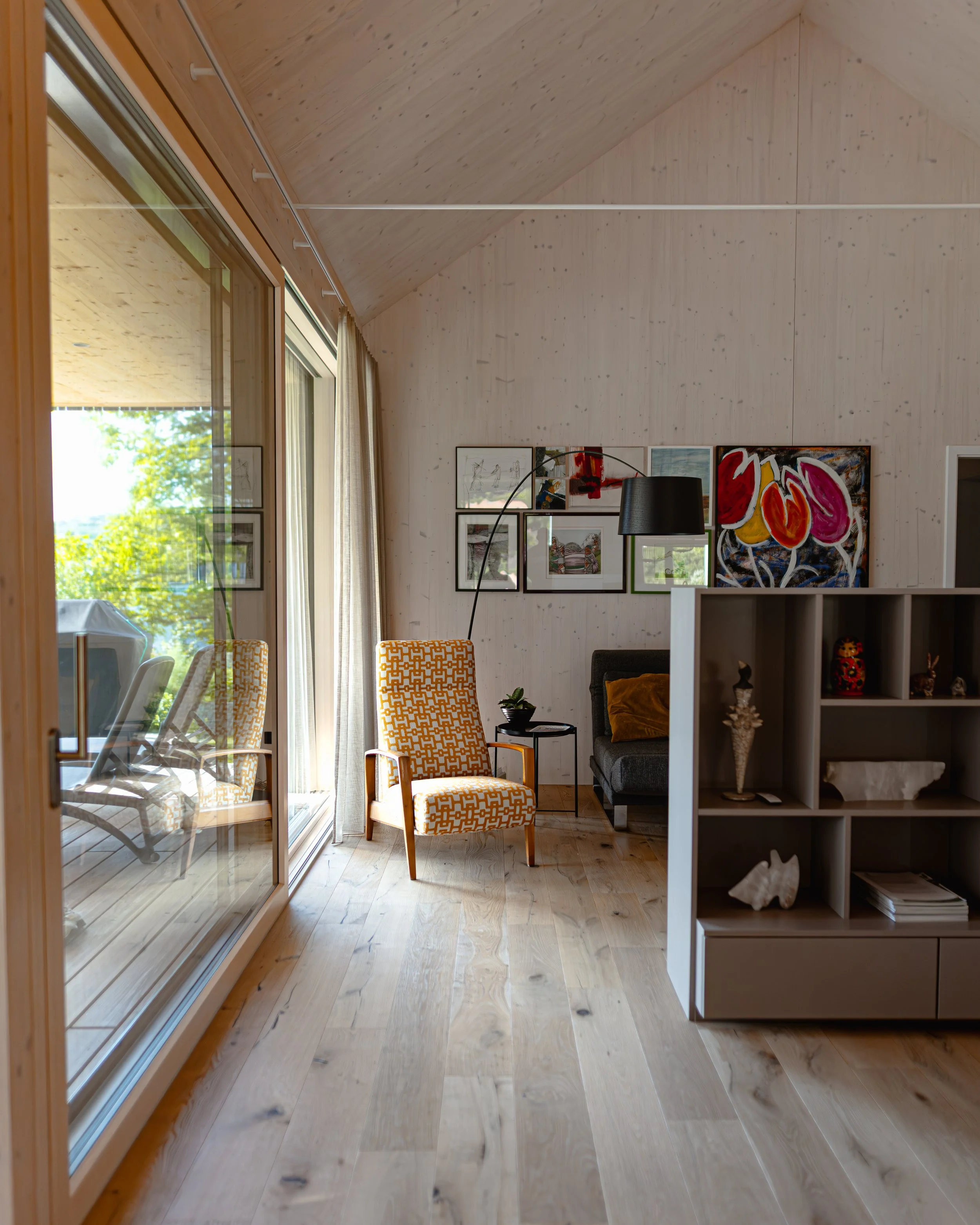 Ein Wohnzimmer mit Holzfußboden, großen Fenstern, gelbem Sessel, grauem Sofa, Stehlampe, Kunstwerken an der Wand und openem Blick auf eine Balkonterrasse im Freien.