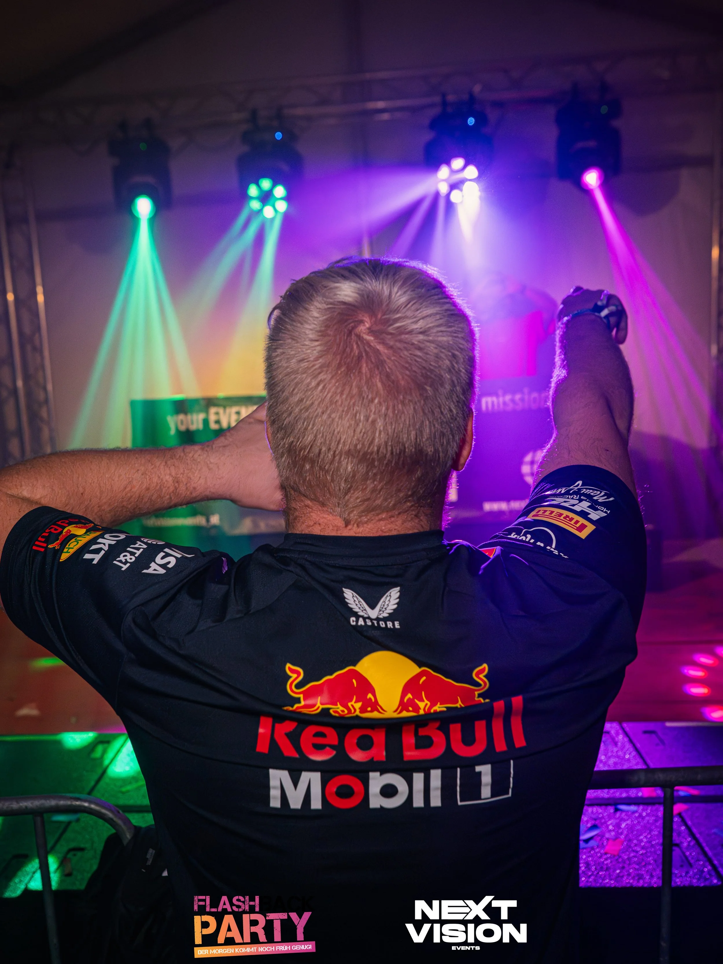 Ein Mann mit einem Red Bull Racing Team T-Shirt schaut auf eine Bühne mit bunter Lichtshow bei einer Partyveranstaltung.