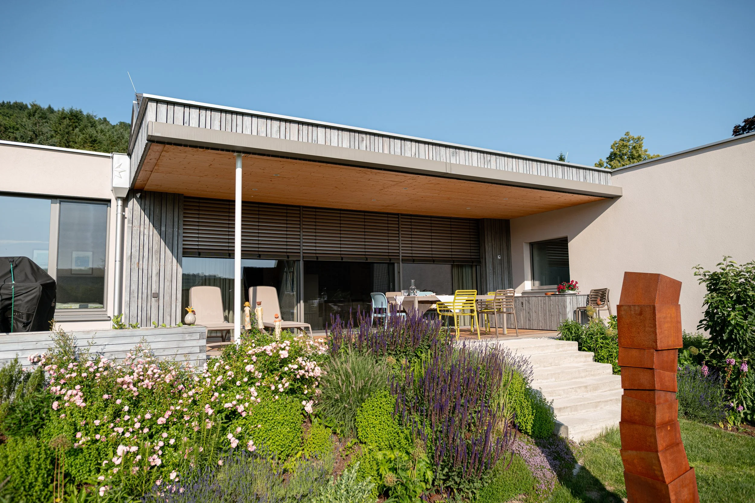 Modernes Haus mit Terrasse, umgeben von Garten mit bunten Blumen und Pflanzen, bei sonnigem Wetter.