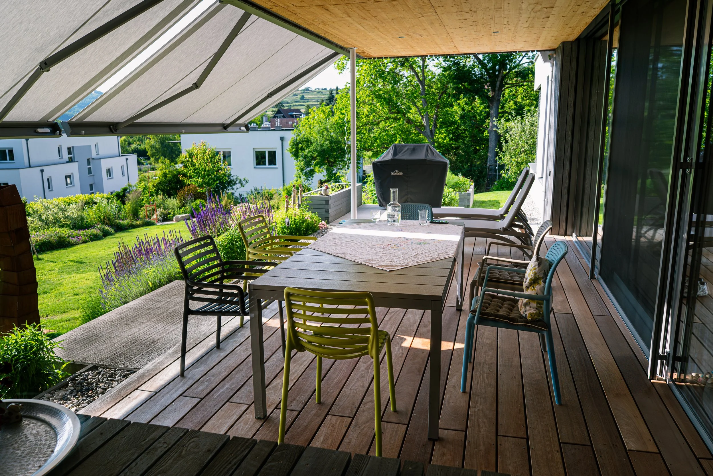 Balkon mit Esstisch, Stühlen, Sonnenliegen und Garten im Hintergrund.
