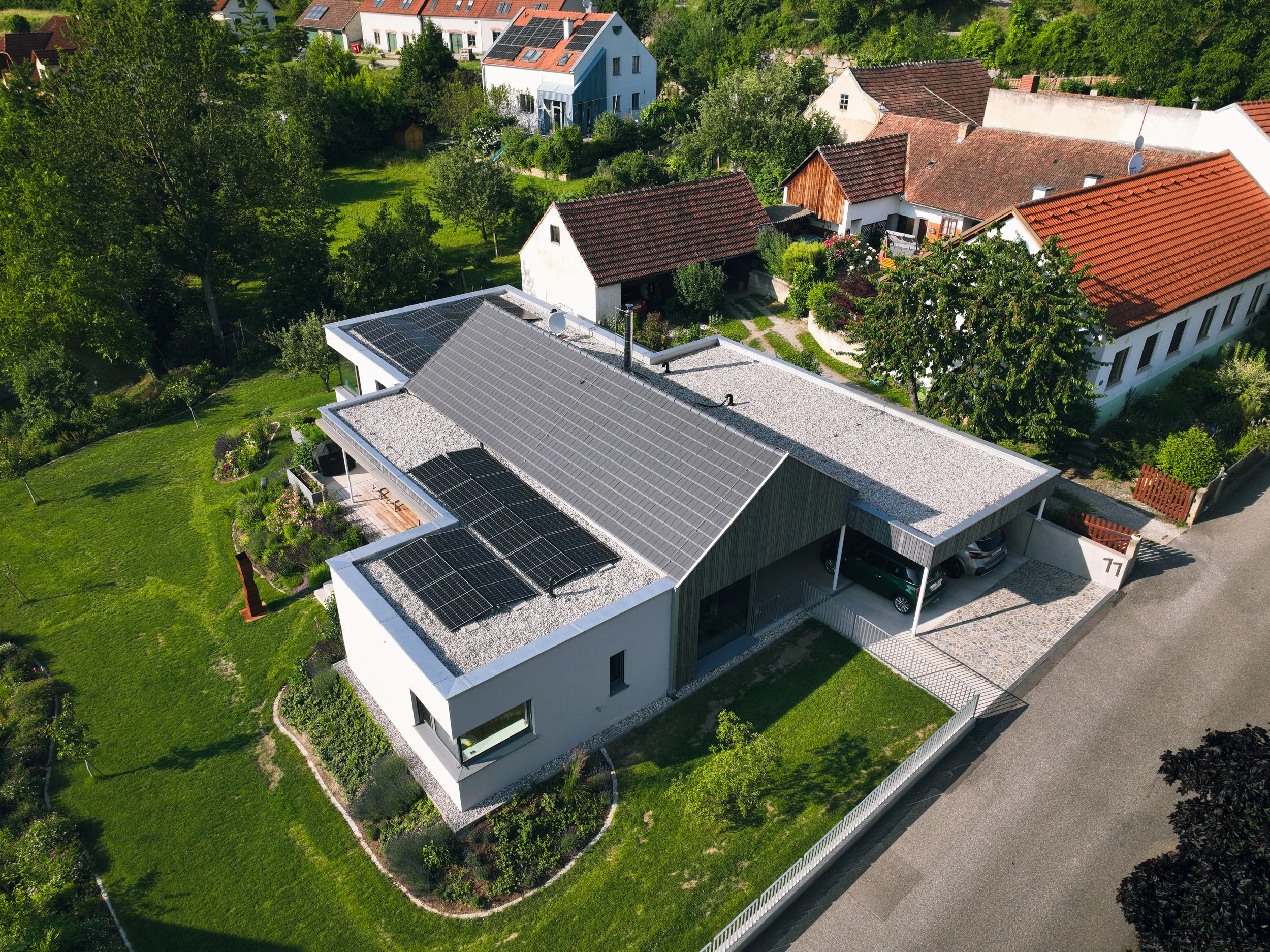 Luftaufnahme eines modernen Hauses mit Solarpanels auf dem Dach, umgeben von einem Garten und anderen Häusern in einer grünen Umgebung.