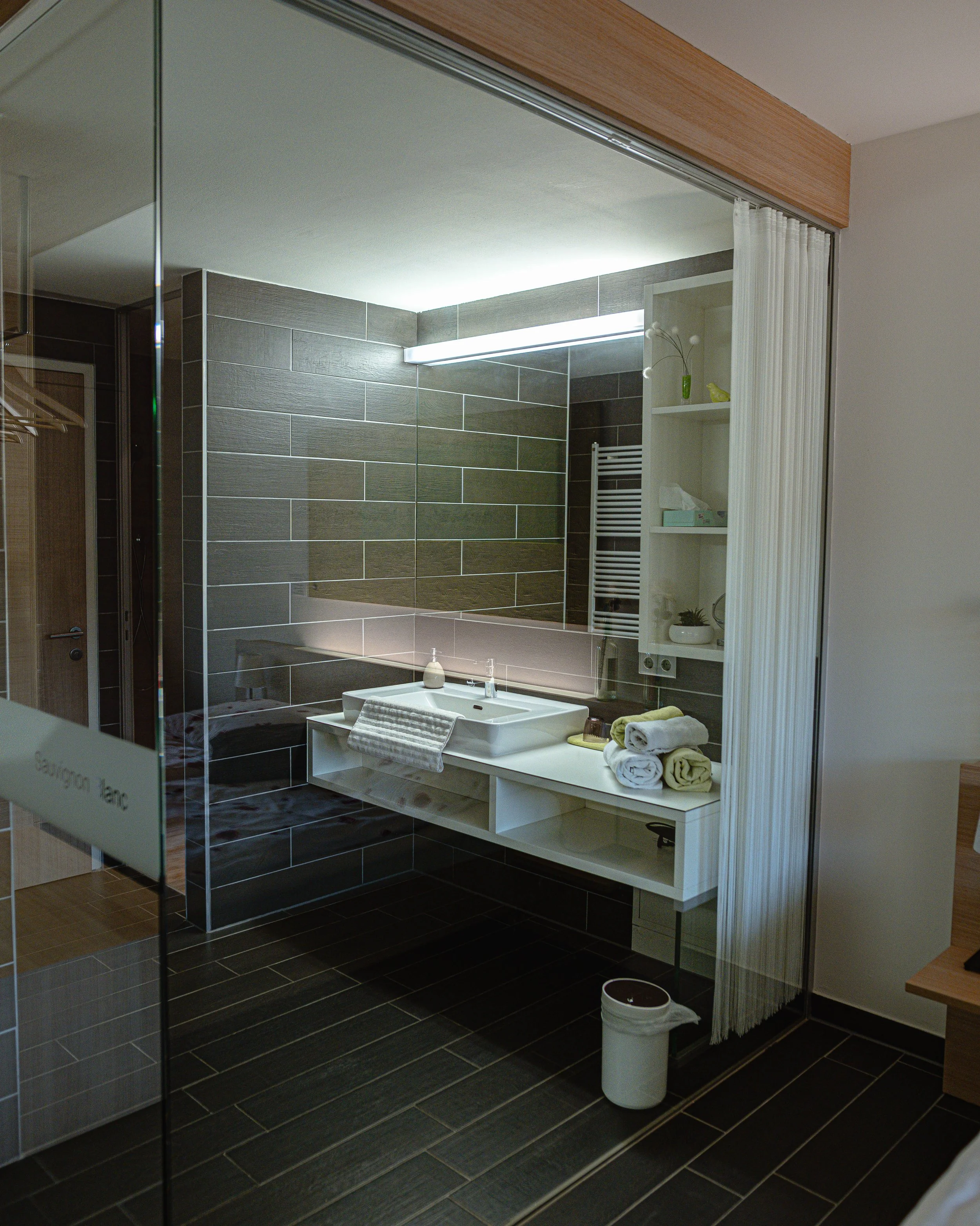 Moderner Badezimmerbereich mit dunklen Fliesen, großem Spiegel, weißen Waschbecken, Handtüchern und Ablageflächen hinter Glasscheibe.