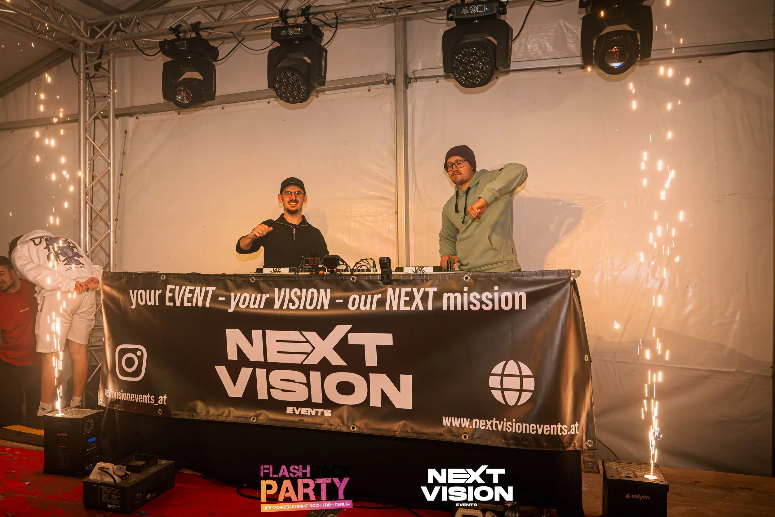 Zwei DJs stehen hinter einem Tisch mit einem Banner, das die Marke 'Next Vision' zeigt, während auf beiden Seiten aufsteigende Funken fliegen. Es ist eine Veranstaltung mit Musik und Beleuchtung.