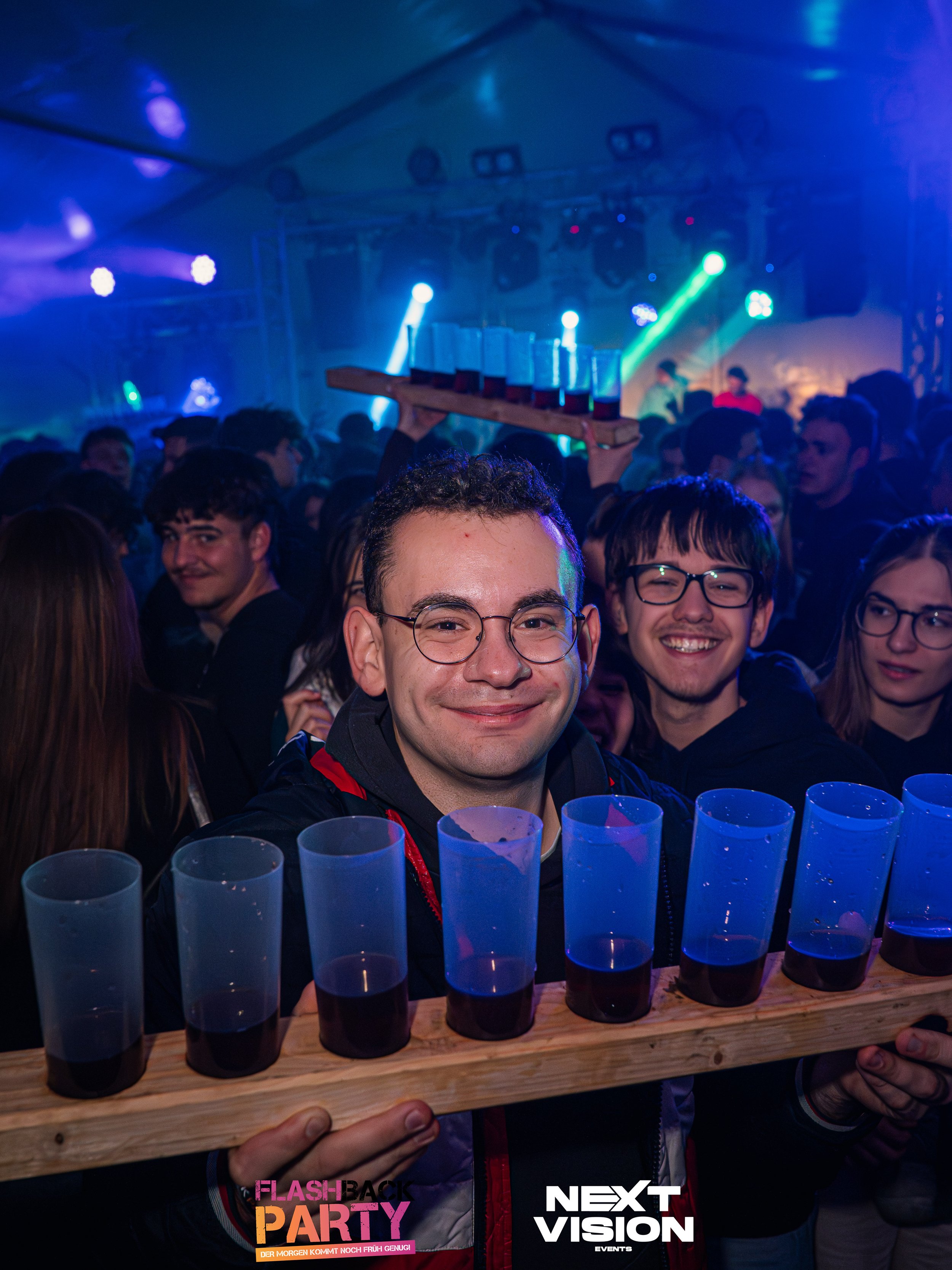 Ein junger Mann mit Brille hält ein Tablett mit mehreren Bechern voller Getränk, umgeben von einer Menschenmenge in einer dunklen Club- oder Party-Atmosphäre mit bunten Lichter.