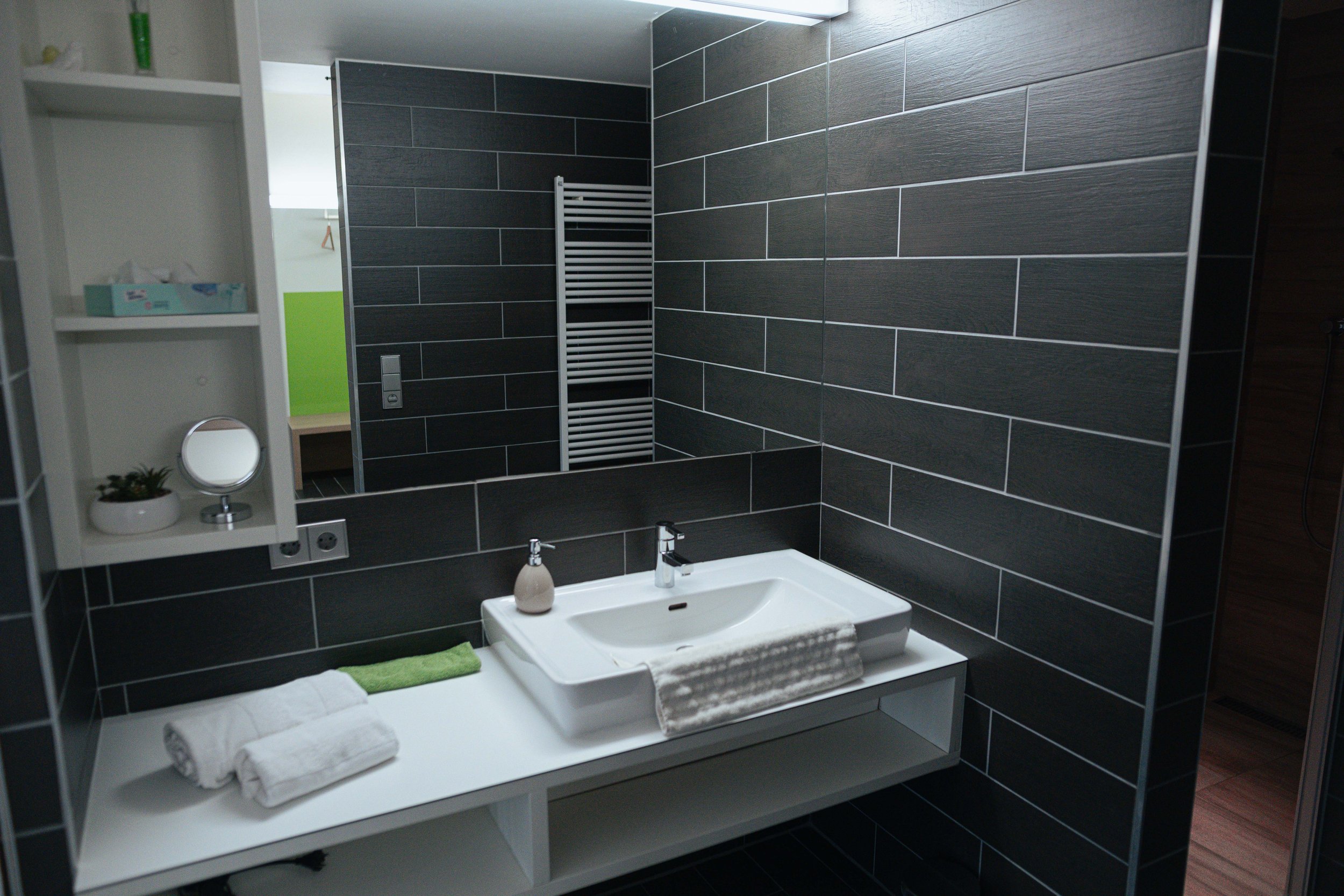 Moderner Badezimmerraum mit schwarzem Wandfliesen, weiße Waschtisch mit Wasserhahn, Handtuch, Seife, Spiegel, weißer Regale, Pflanzen, und Heizkörper im Hintergrund.