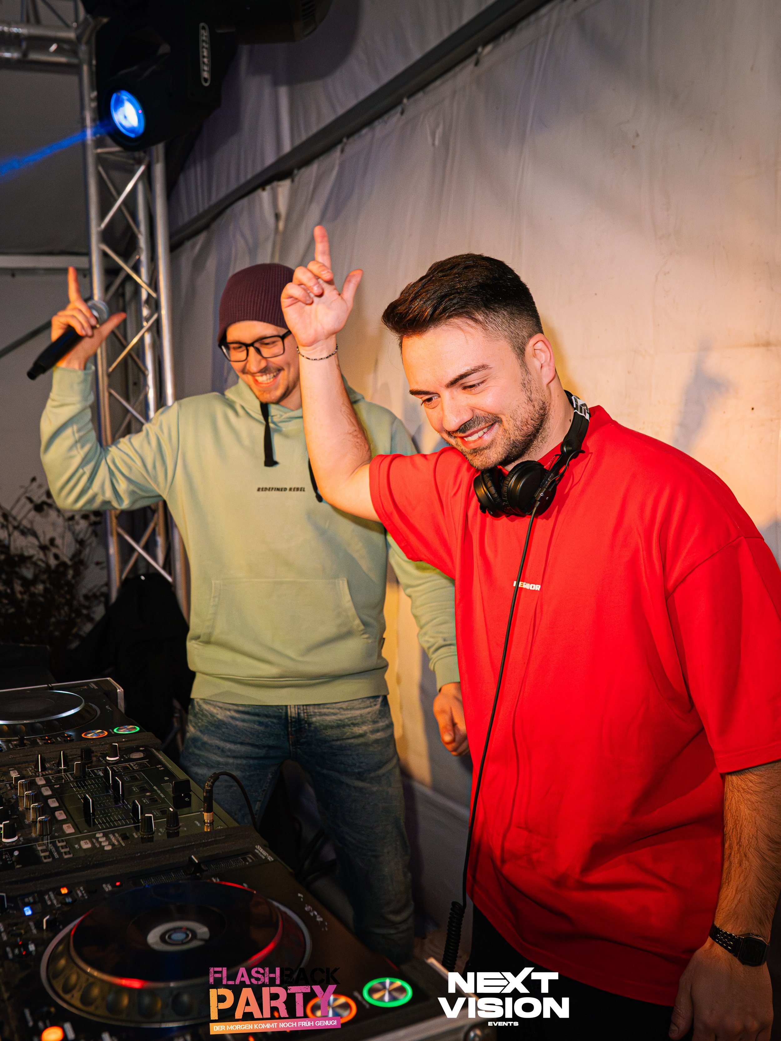 Zwei Männer beim Auflegen bei einer Party, die vor einem DJ-Setup stehen und tanzen. Einer trägt eine grüne Kapuzenjacke und eine Brille, der andere trägt ein rotes T-Shirt und Kopfhörer. Im Hintergrund ist ein Beleuchtungsgerät.