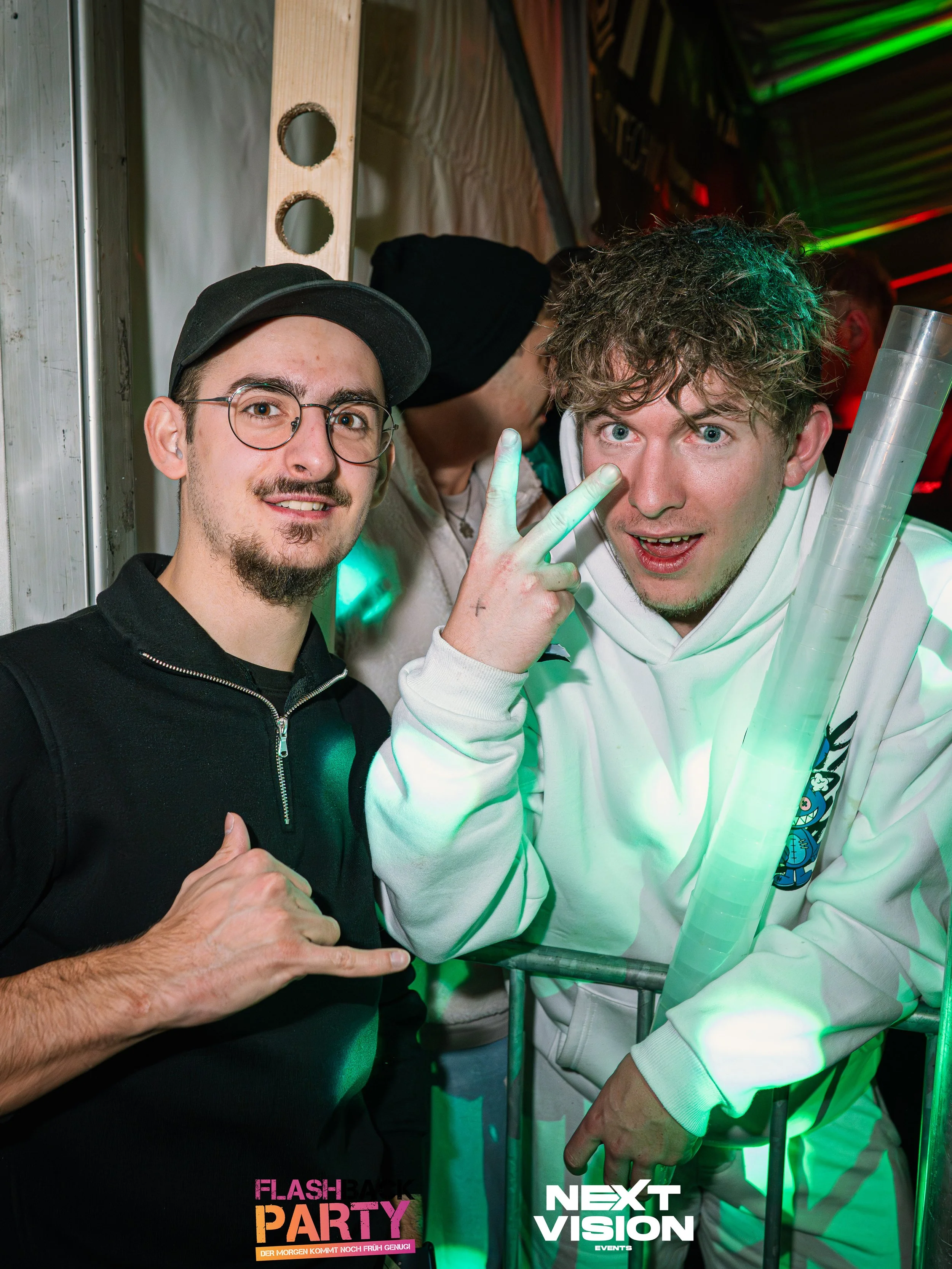 Zwei junge Männer posieren auf einer Party. Einer trägt eine schwarze Jacke und eine Kappe, der andere eine weiße Hoodie, der den Arm mit Leuchtstäben hält. Beide schauen in die Kamera, der Mann in Weiß zeigt eine Peace-Geste, beide scheinen Spaß zu 