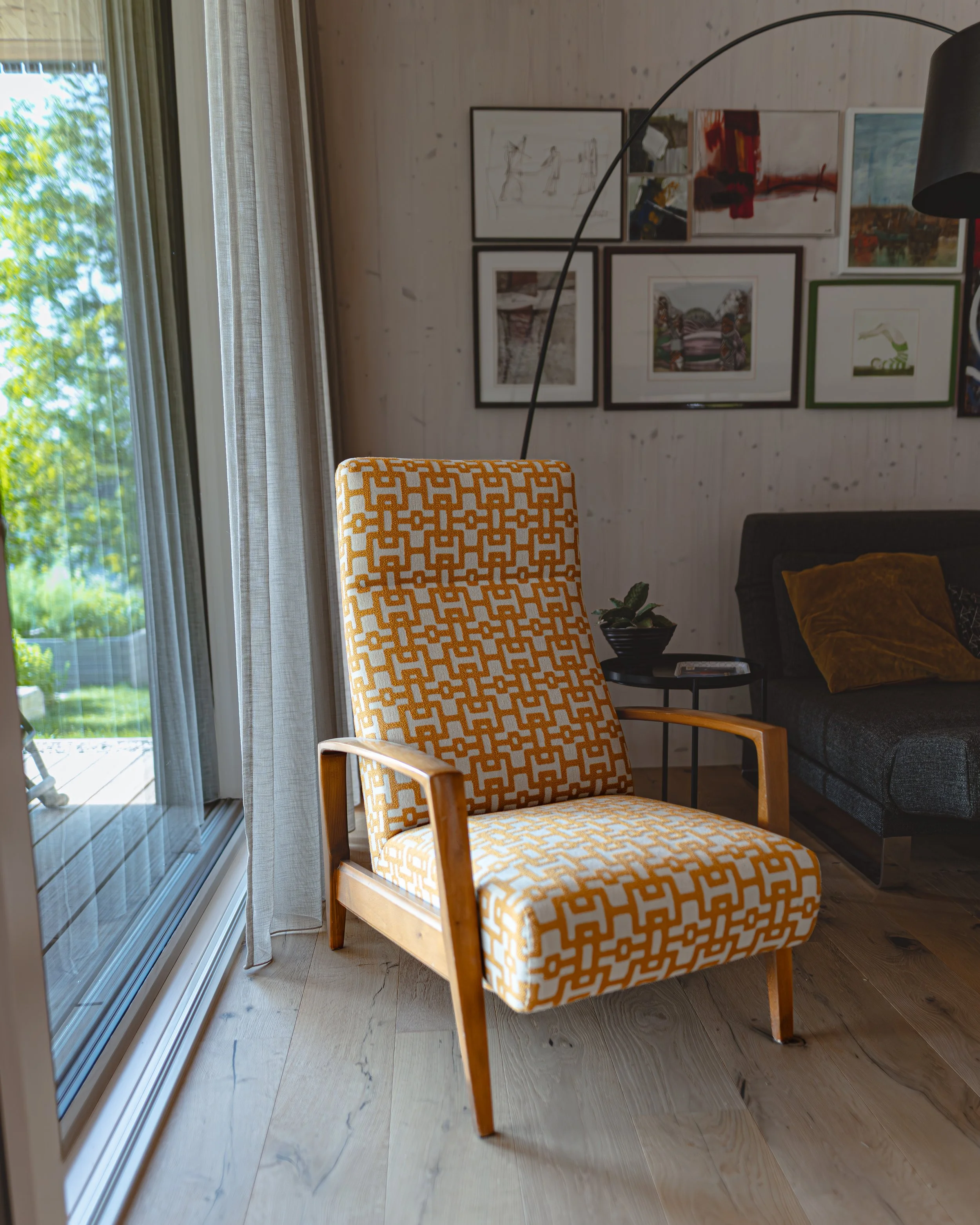 Sitzgelegenheit mit orange-weiß gemustertem Stoff in einem modernen Wohnzimmer, Fenster mit Vorhängen, Wanddekoration mit mehreren Bilderrahmen und Couch mit Kissen.