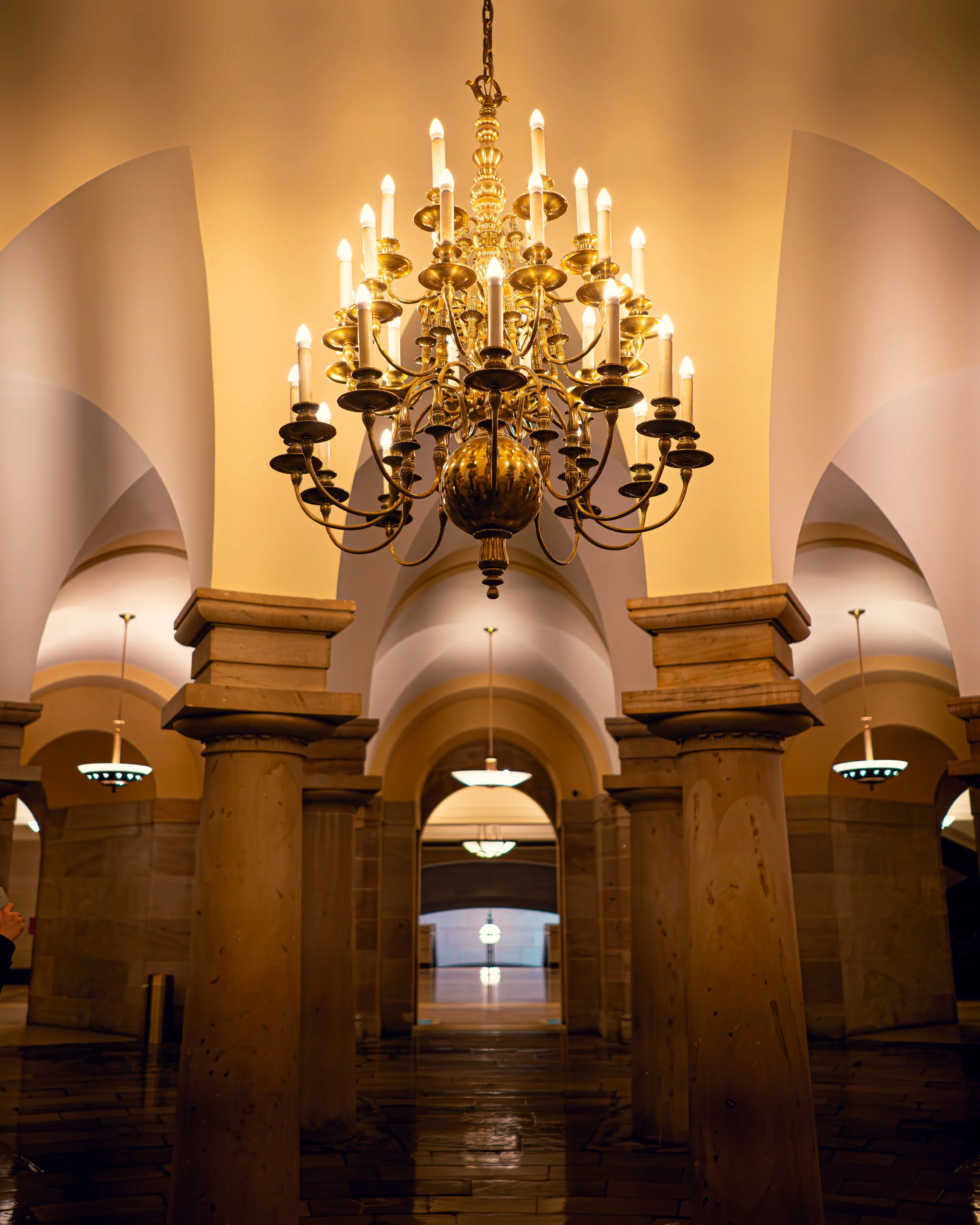 Ein prachtvoller, goldener Kronleuchter hängt von der Decke, beleuchtet einen eleganten Raum mit weiß gewölbten Decken und großen runden Säulen, die den Raum stützen.