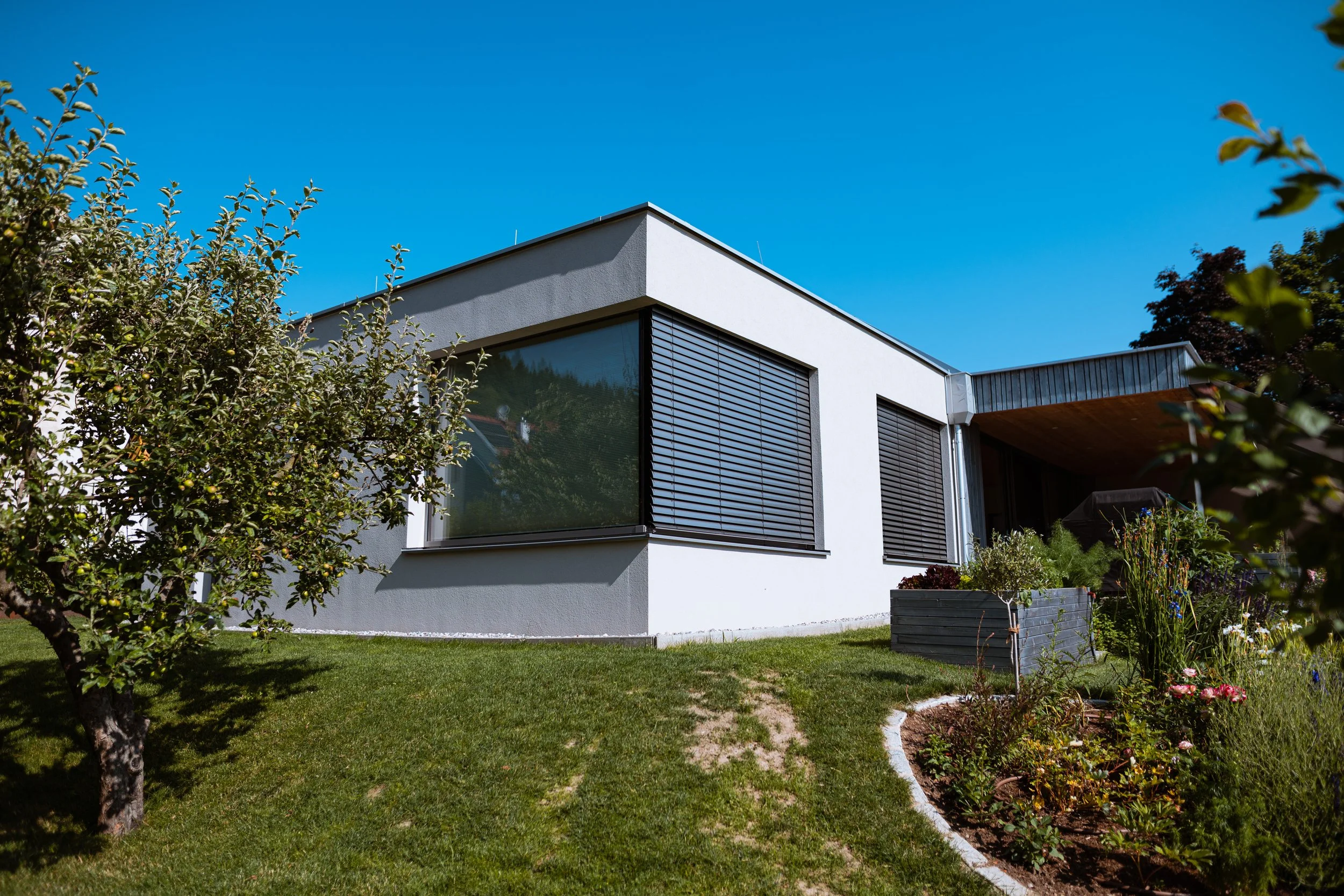 Modernes weißes Haus mit großen Fenstern und Rollläden, umgeben von einem gepflegten Garten mit Bäumen und Blumen, unter klarem blauen Himmel.