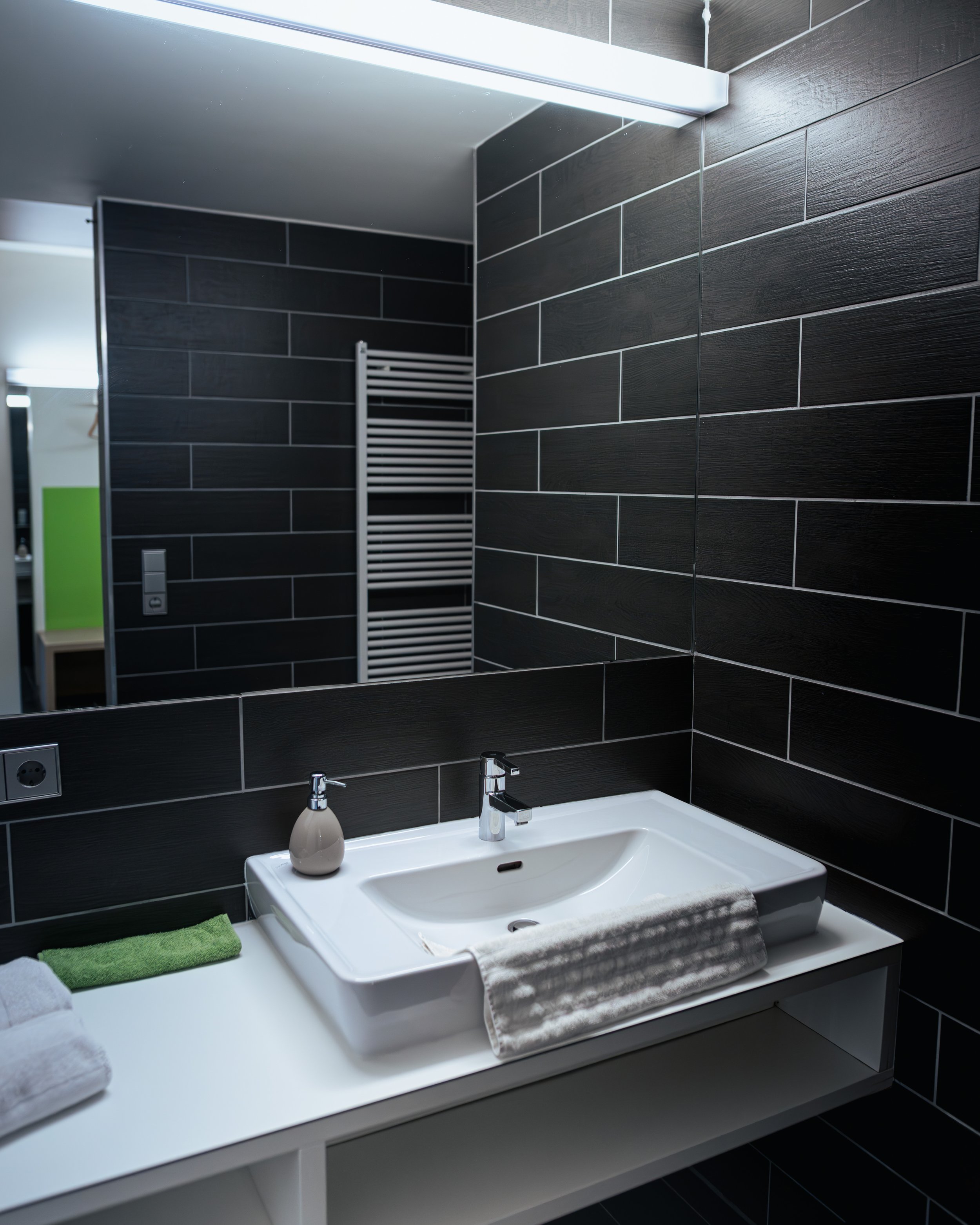 Moderne schwarze Badezimmerfliesen, Waschtisch mit weißen Handtüchern, Hamamelis-Soap und Wasserhahn, großer Spiegel, beleuchtete Decke, Heizkörper im Hintergrund.