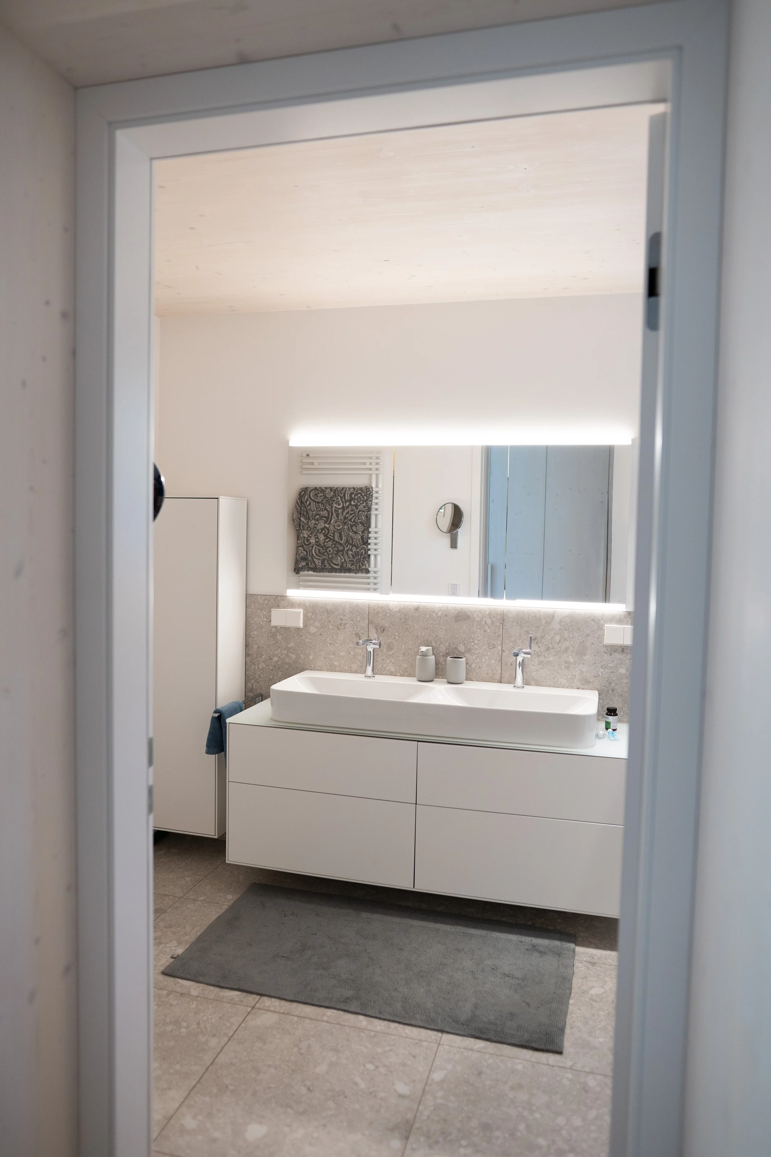 Moderner Badezimmerblick durch Tür auf Doppelwaschbecken mit großem Spiegel und Handtuchhalter in Hintergrund.