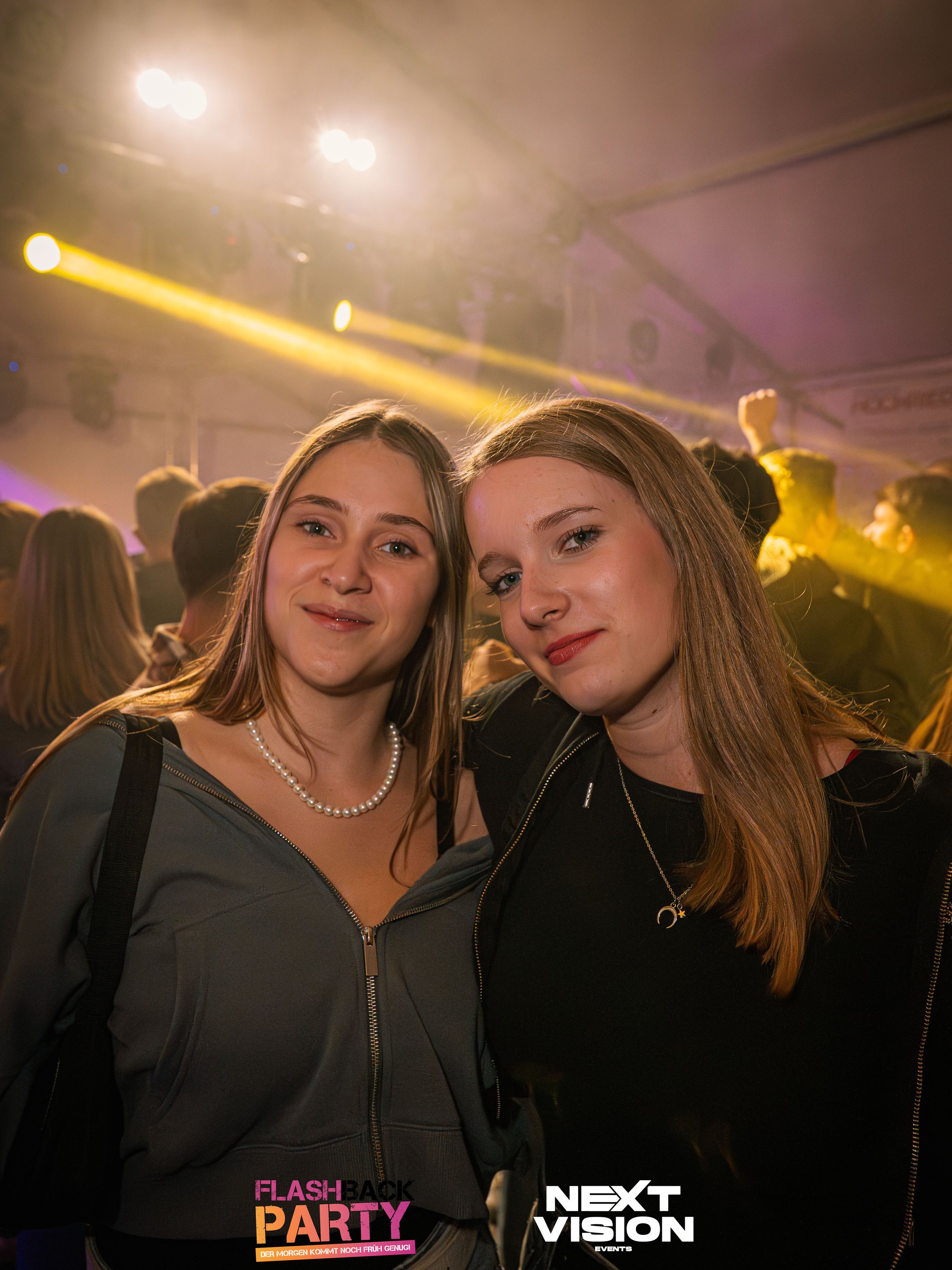 Zwei junge Frauen beim Feiern auf einer Party, lachend in einer Tanzveranstaltung mit bunten Lichtern im Hintergrund.