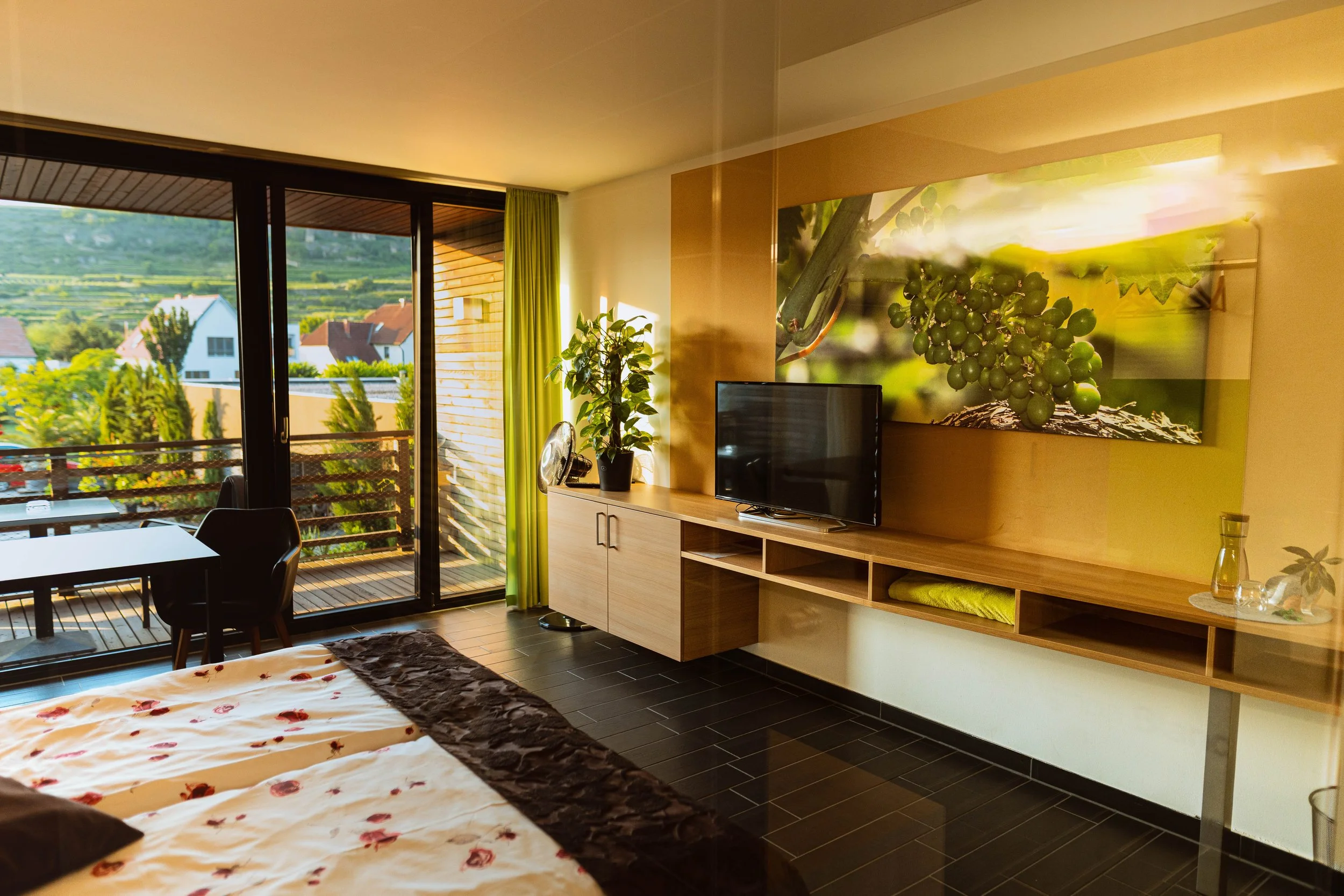 Schlafzimmer mit Balkon, TV, Pflanzen, Wandbild von Weintrauben, Bett mit floral bedruckter Decke, dunkler Boden, große Fenster mit grünen Vorhängen, Deko-Objekte wie Wasserkrug und Kerze.