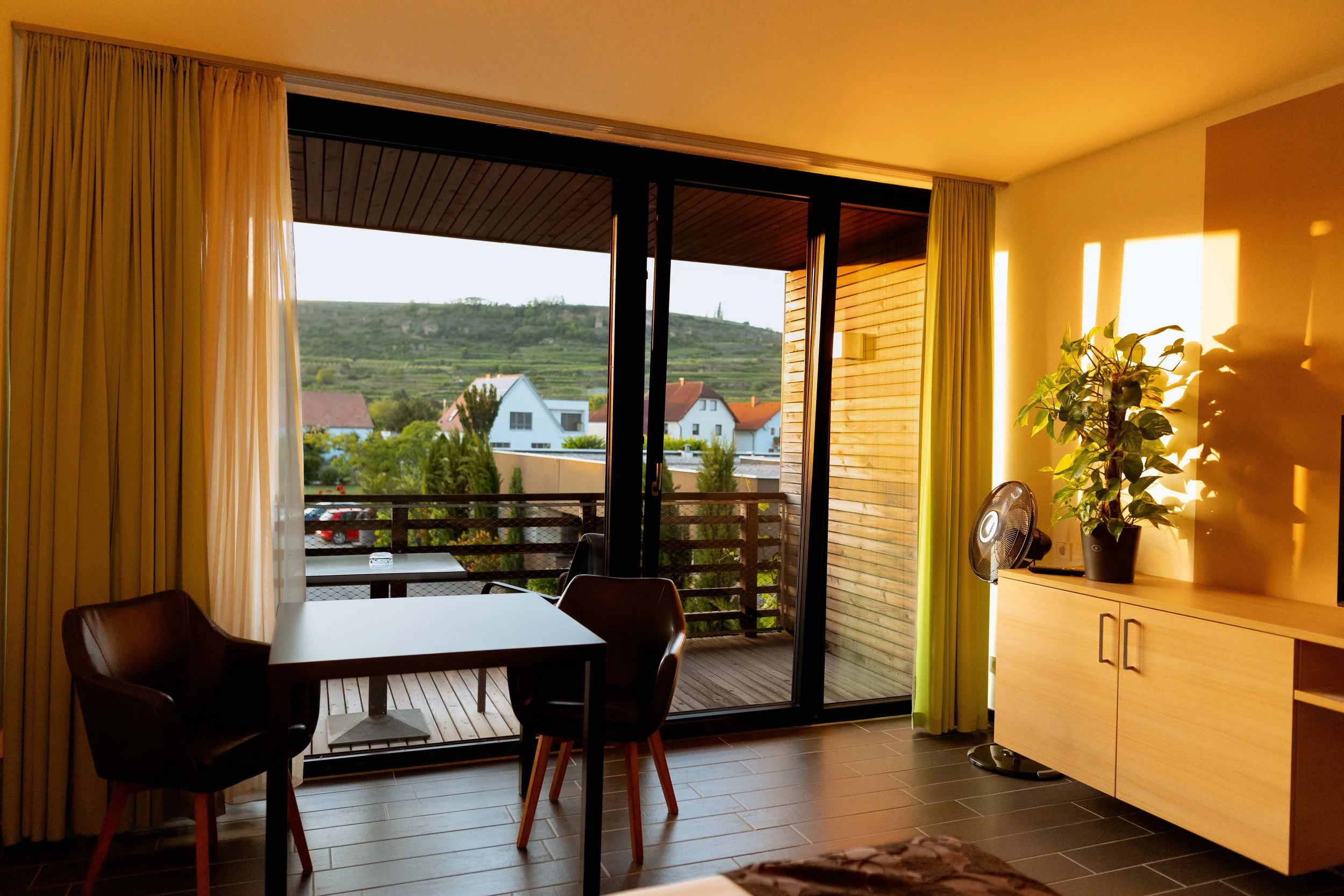 Innenraum eines modernen Zimmers mit einem Glas-Schiebefenster, das auf einen Balkon mit Holzboden führt, und einer Aussicht auf Häuser im Grünen im Hintergrund.