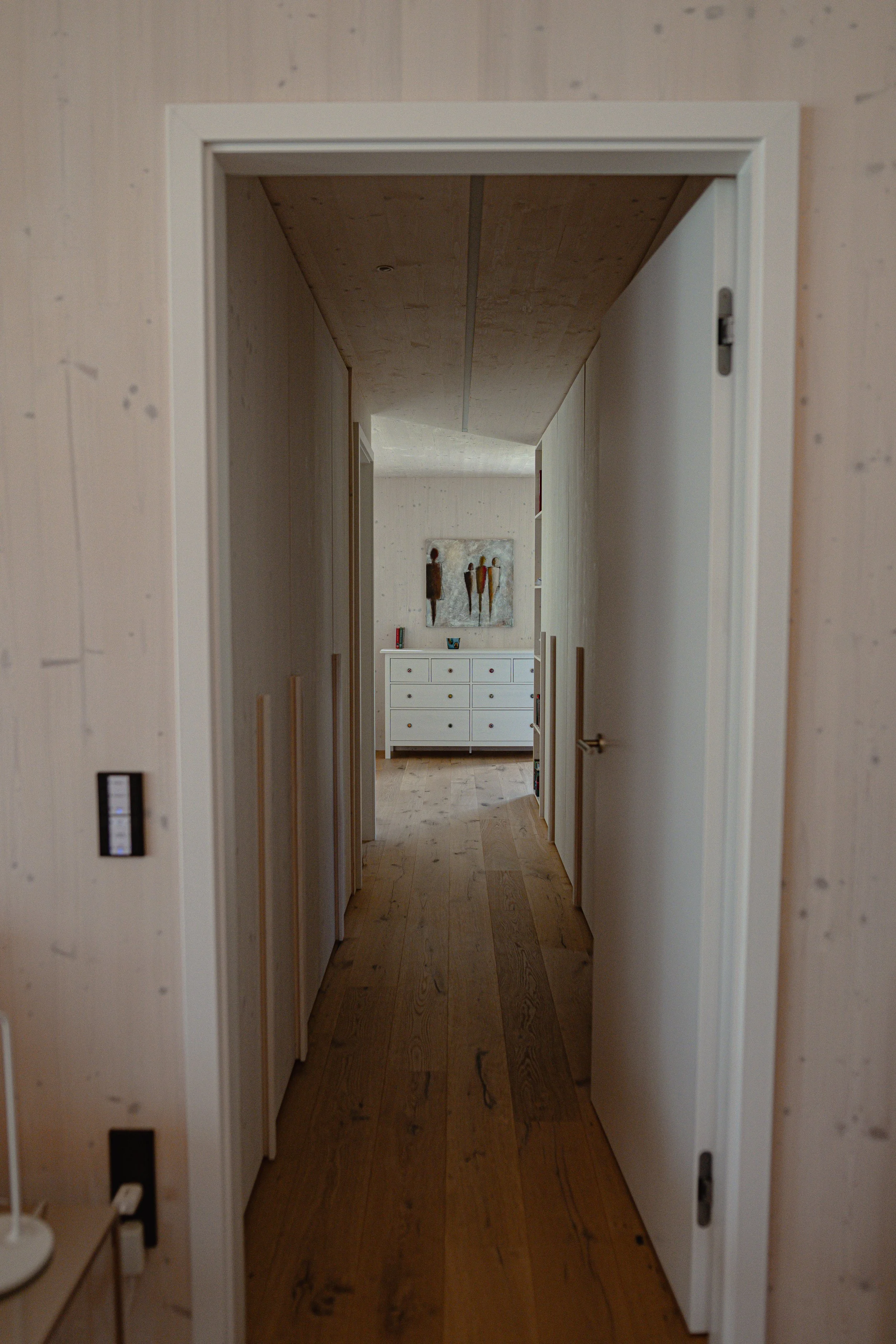 Ein heller Flur in einem Haus mit Holzboden und weißen Wänden, am Ende befindet sich eine weiße Kommode mit mehreren Schubladen, darüber hängt eine Wanddekoration mit drei stilisierten Figuren.