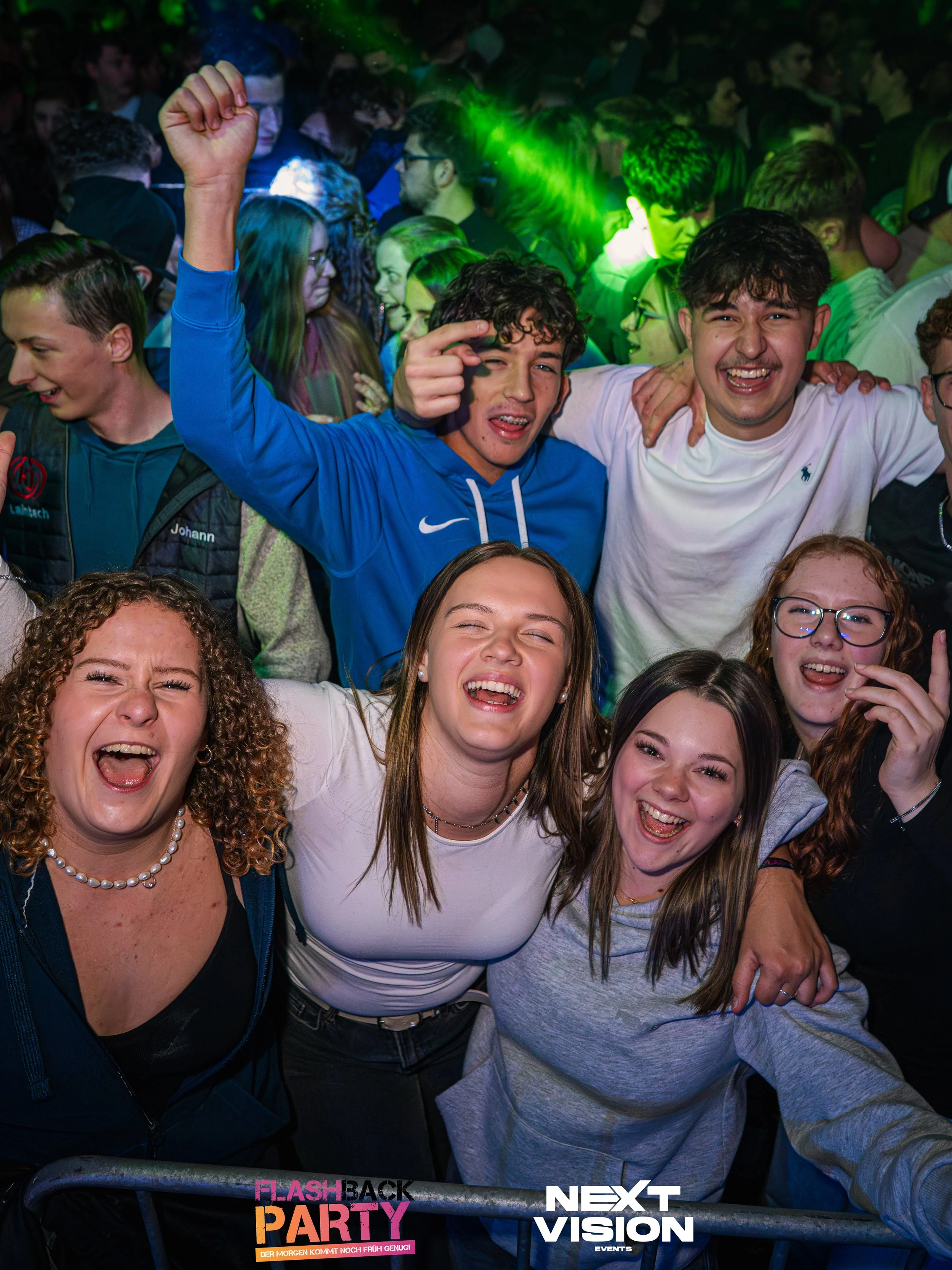 Eine Gruppe lachender und tanzender Jugendlicher bei einer Party, mit Lichteffekten und einer Barriere im Vordergrund, auf der das Logo ‚Flashback Party‘ und ‚Next Vision Events‘ zu sehen sind.