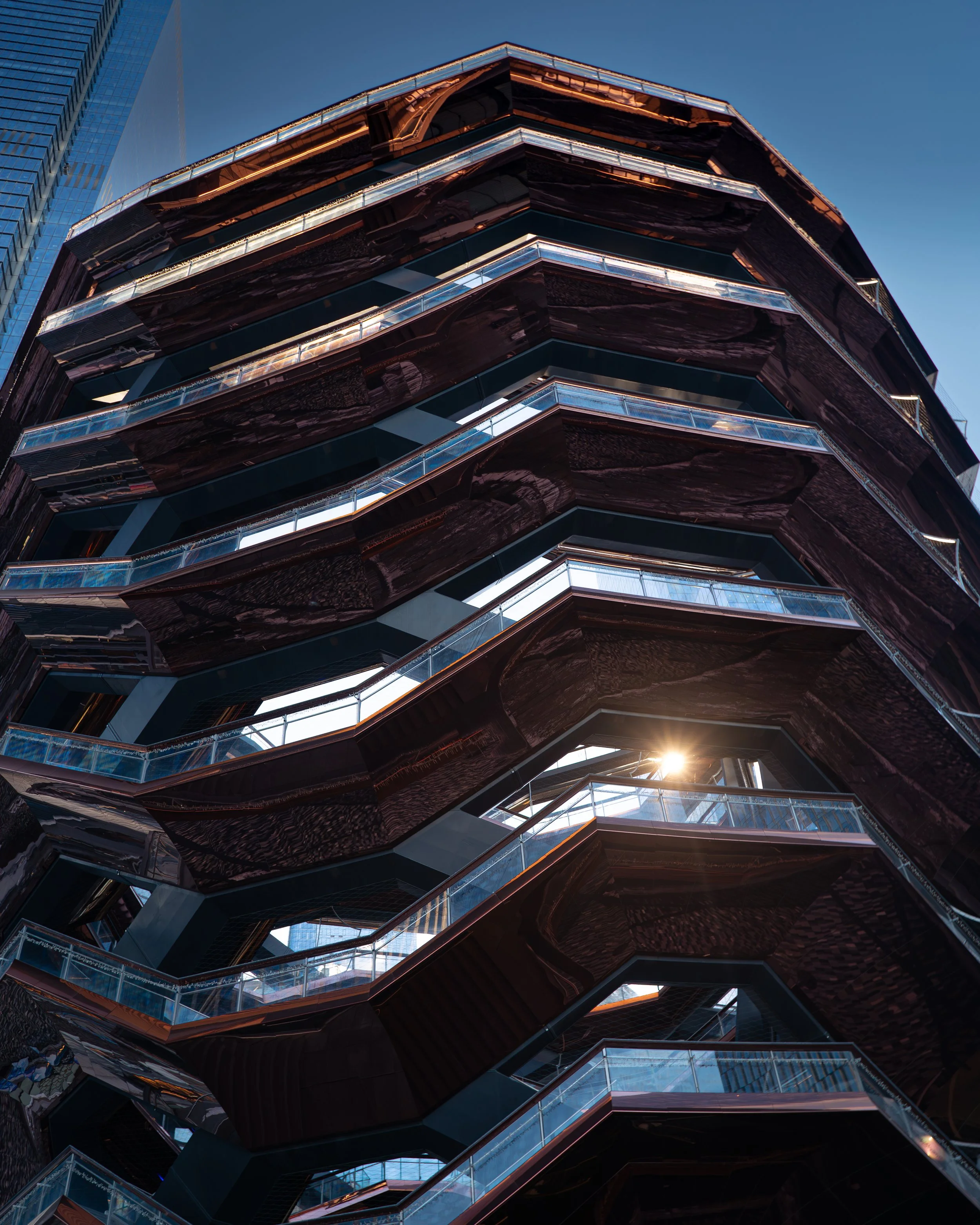 Nahaufnahme eines modernen, mehrstöckigen Gebäudes mit dunklen, gewundenen Fassadenelementen und Glasbrüstungen, bei Sonnenuntergang oder -aufgang.
